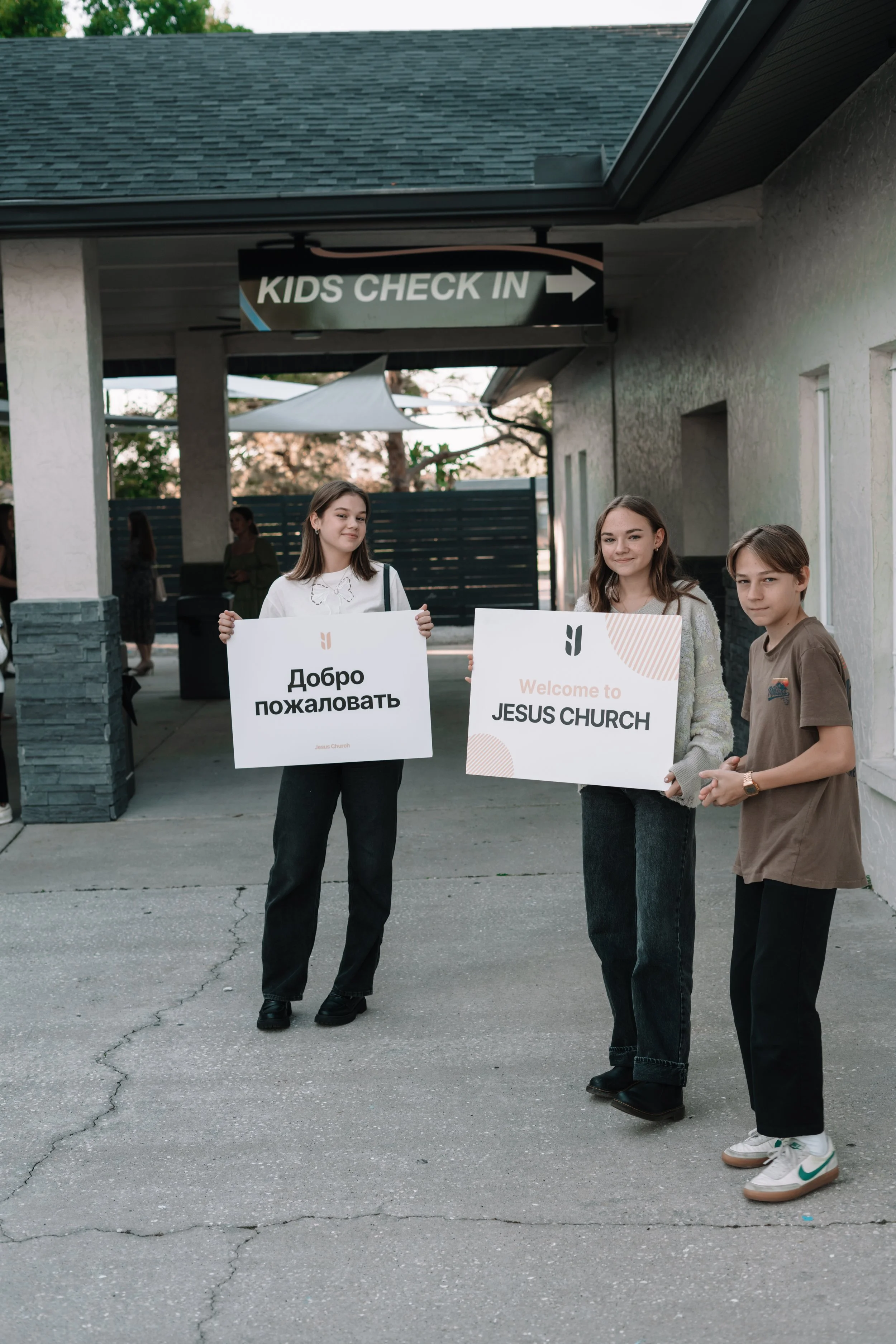 Three teenagers standing outside a church holding signs welcoming visitors, with a sign overhead reading 'Kids Check In' and a fence in the background.