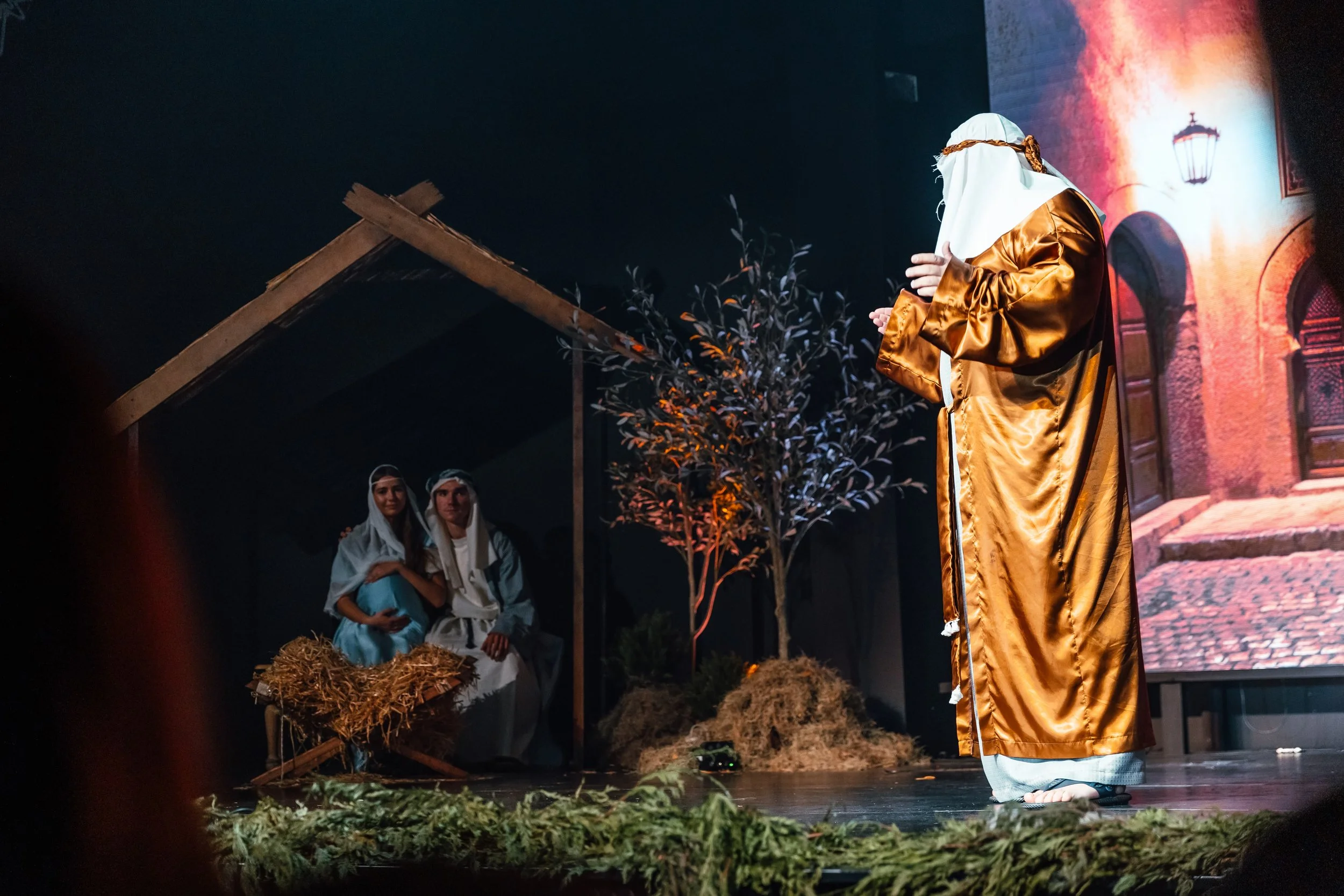 Performers engaged in a nativity scene play on stage, depicting a shepherd wearing a gold robe, two women sitting under a shelter with a manger, a wooden stable structure, and a backdrop resembling a Mediterranean village with a lamp post.