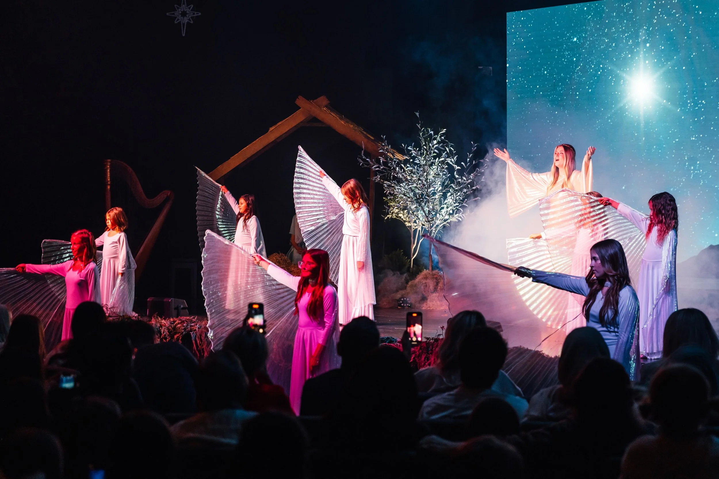 Performers on stage dressed in white robes, holding large fan-like props, with a backdrop of a starry sky and dusk scenery, while an audience watches, some taking photos.