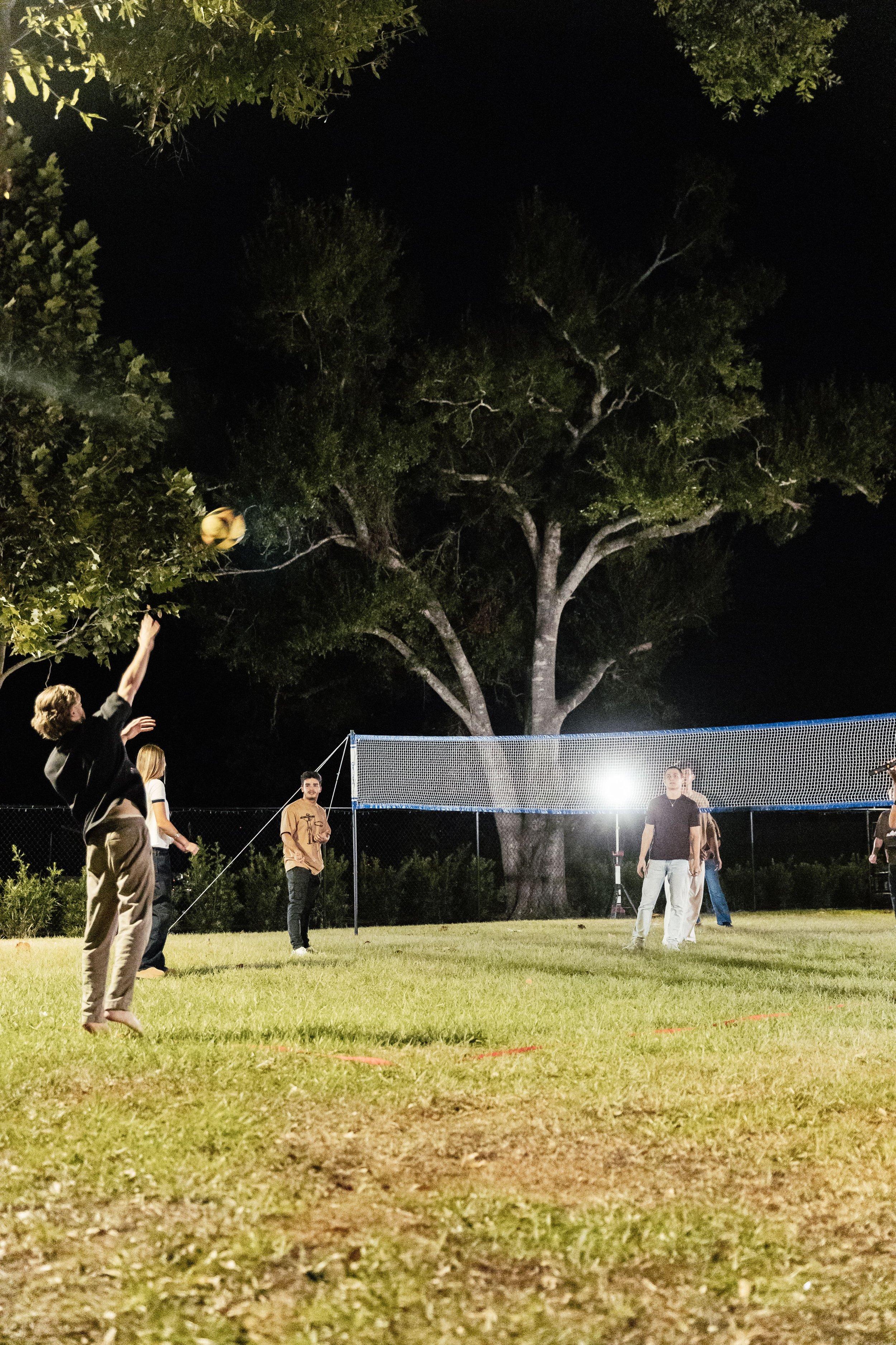 People playing volleyball outdoors at night on a grassy field with large trees in the background.