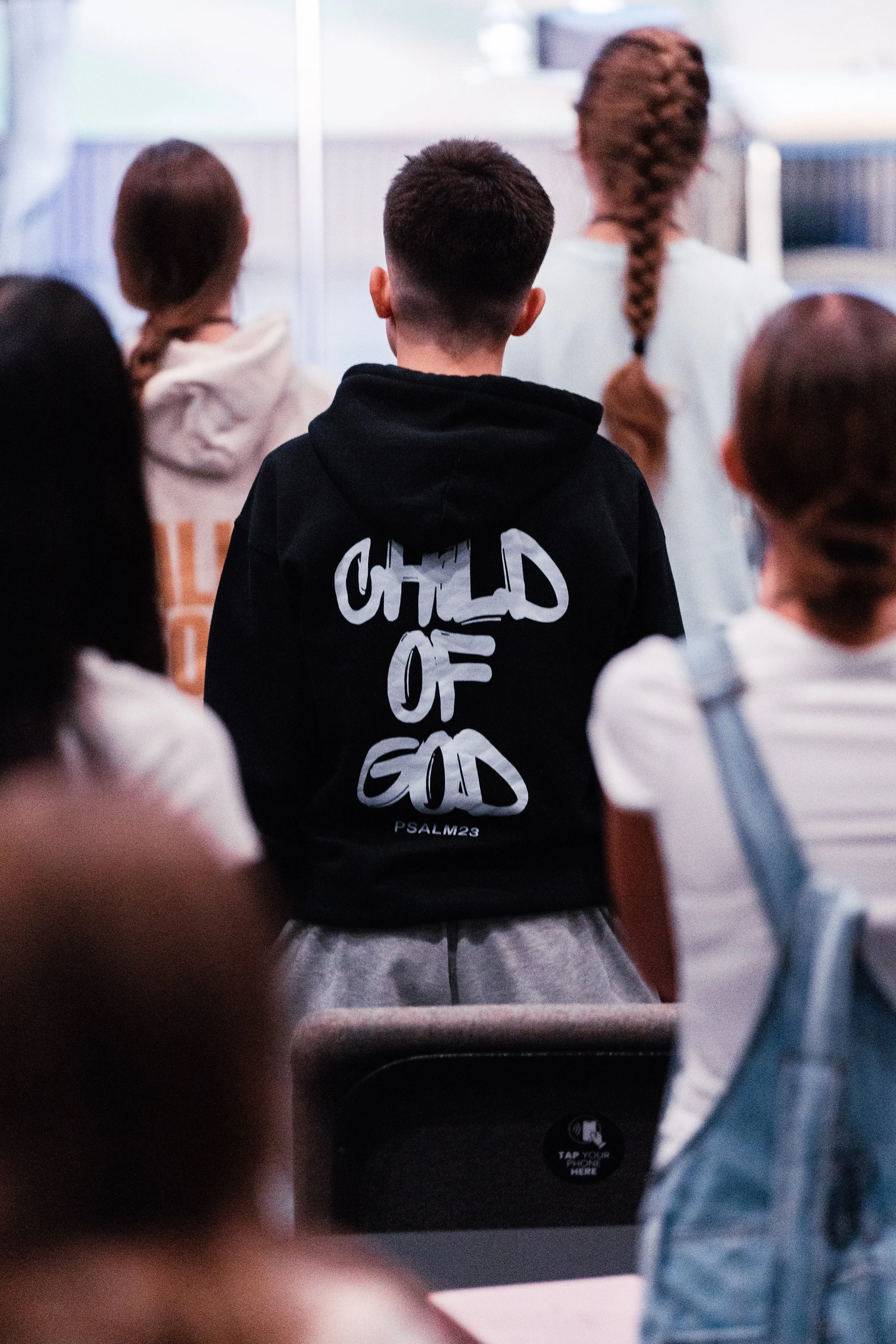 A group of children sitting in a room, with one boy in a black hoodie with 'Children of God' written on the back, facing forward.