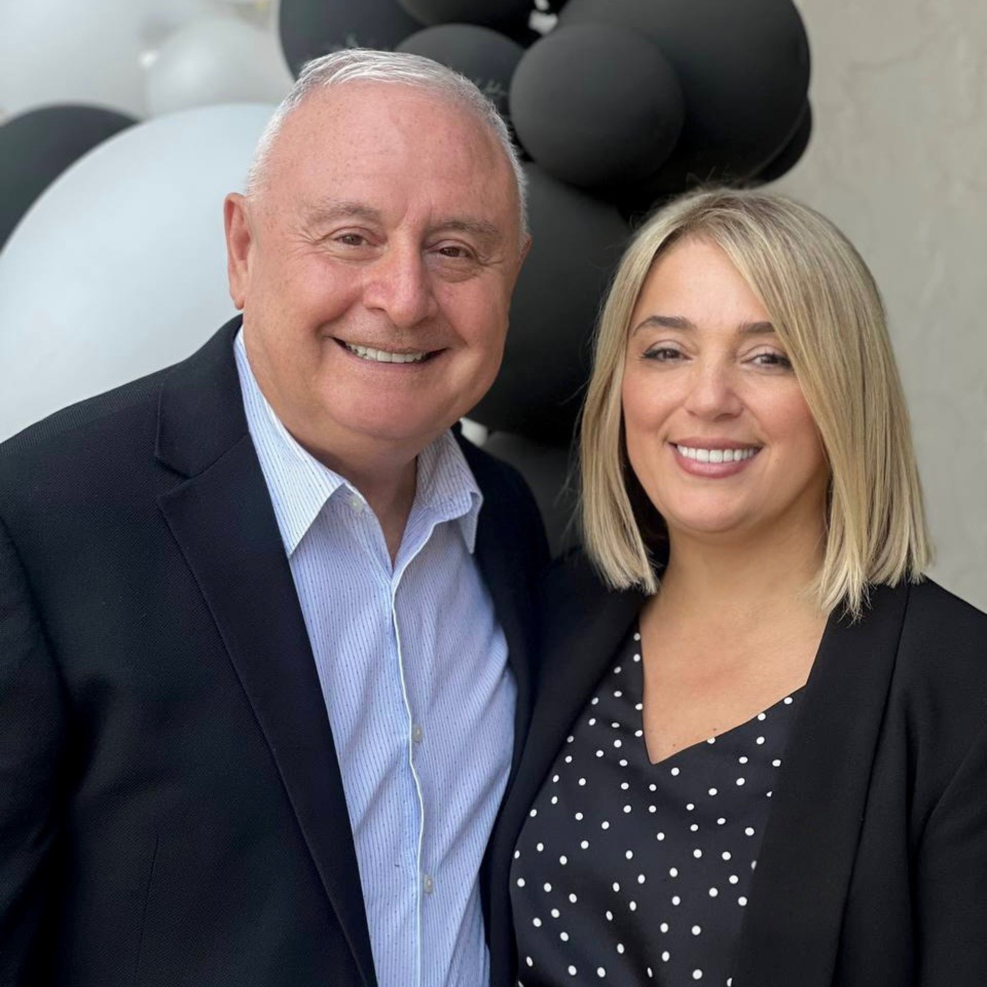A smiling man and woman pose together in front of black and white balloons. The man is in a dark suit and a light striped shirt, and the woman is in a black blazer over a polka dot blouse.