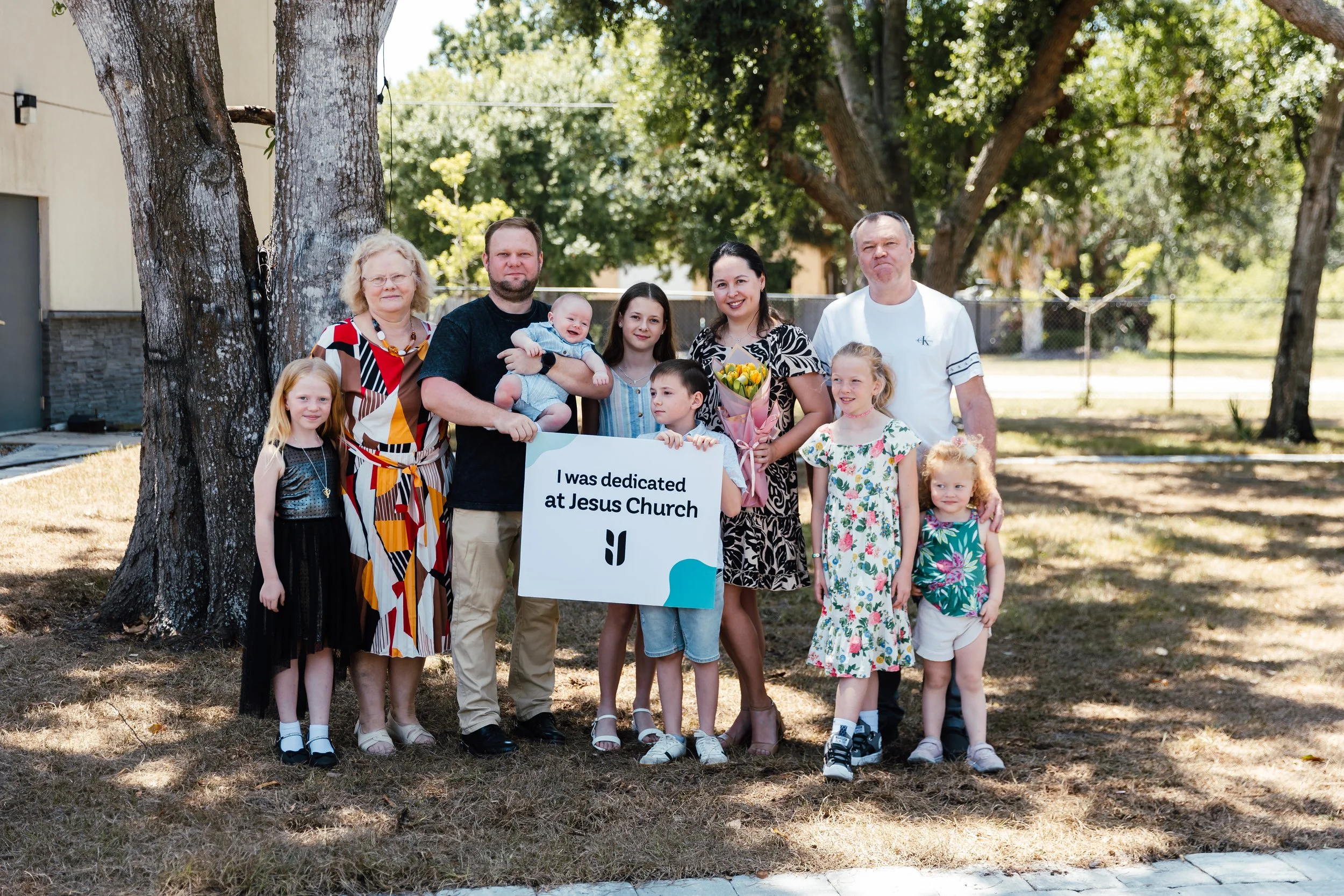 Family group standing outdoors near a large tree, holding a sign that reads 'I was dedicated at Jesus Church,' with a woman holding a bouquet of flowers and children of various ages surrounding them.