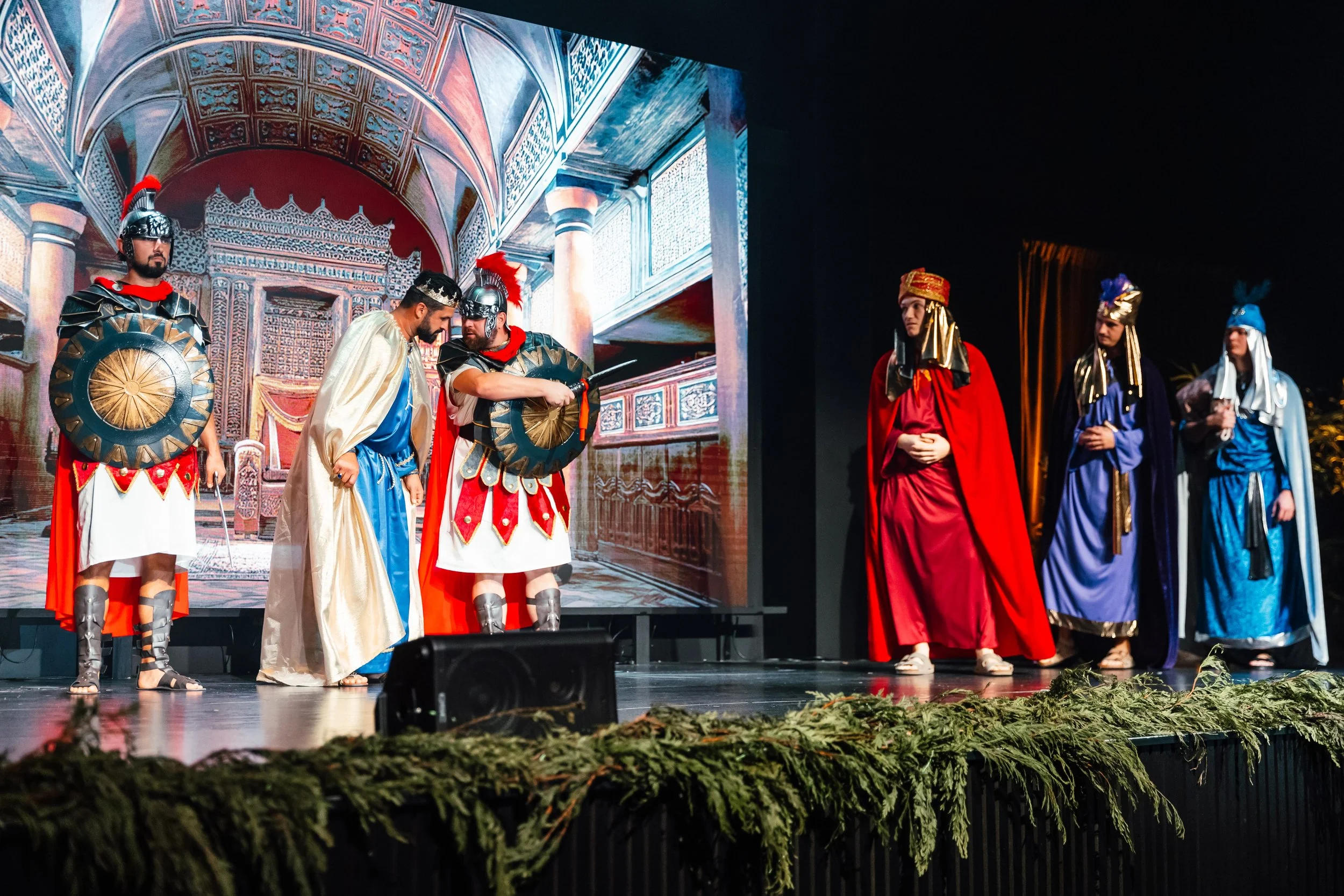 Actors dressed as Roman soldiers and historical figures performing a theatrical play on stage with a digital backdrop depicting a Roman palace.