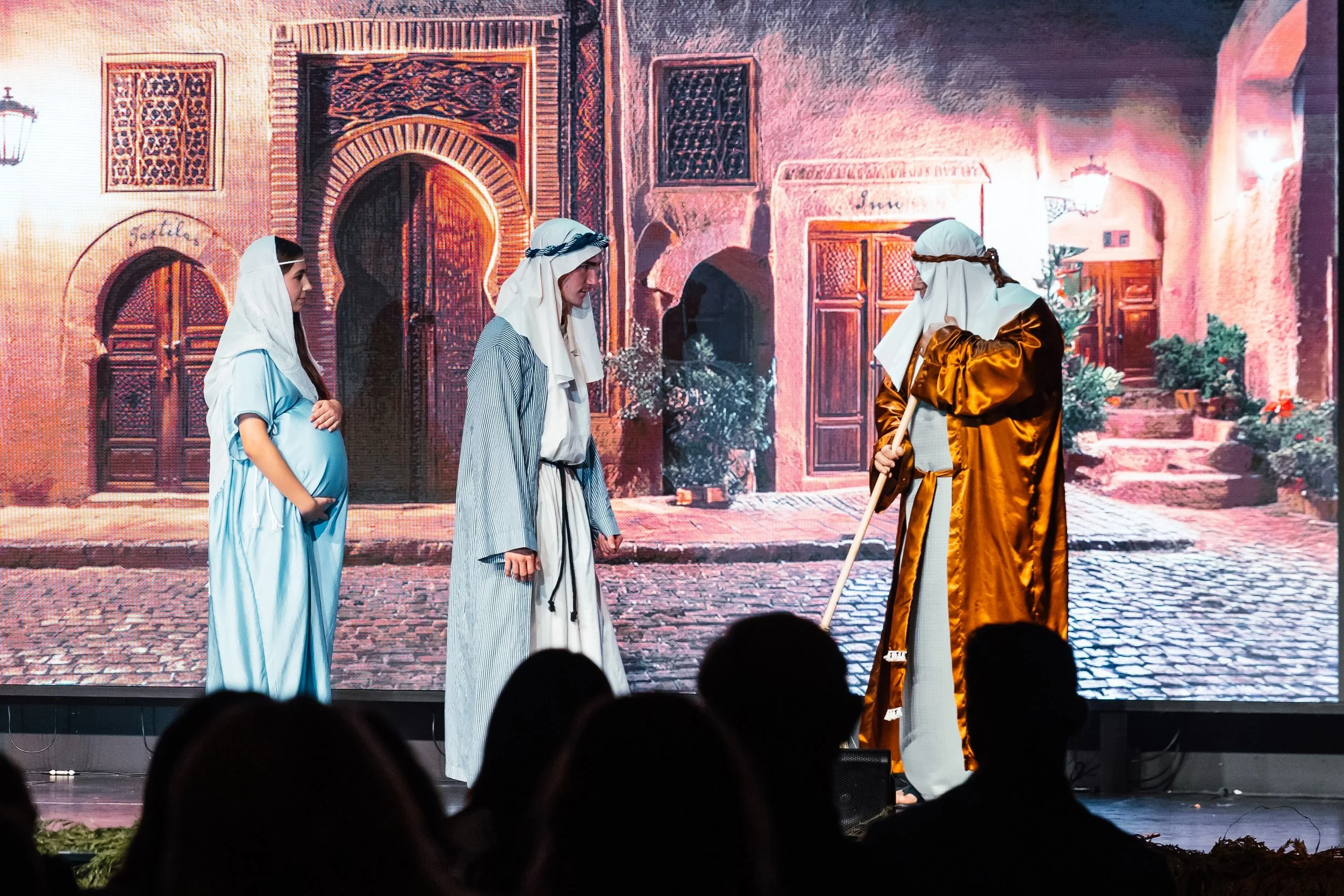 A nativity scene performance with four actors dressed as biblical figures, standing on stage in front of a backdrop depicting an old town street, with audience members visible in the foreground.