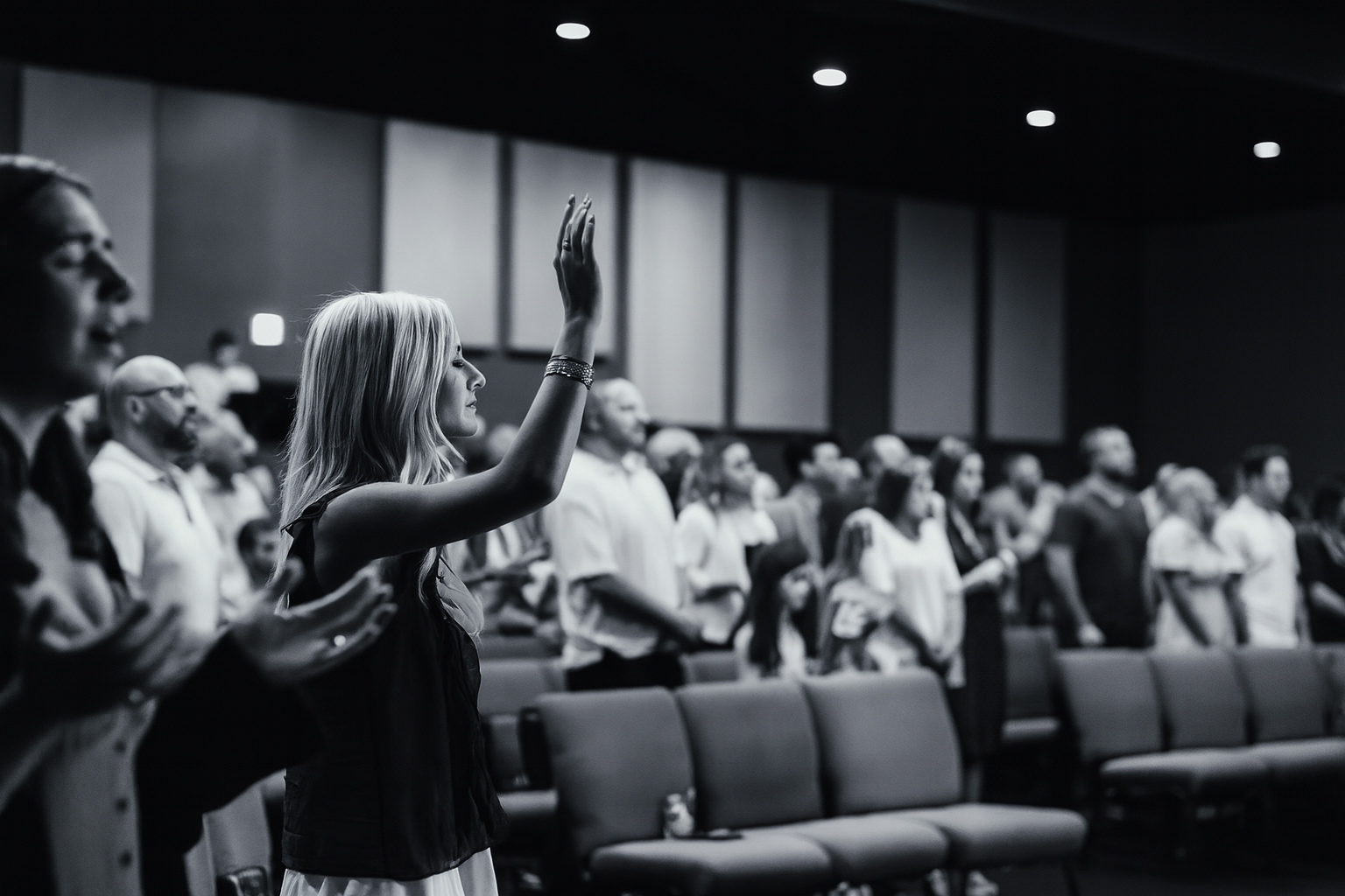 A woman raising her right hand during a prayer or pledge in a church or conference hall with other people standing and participating.