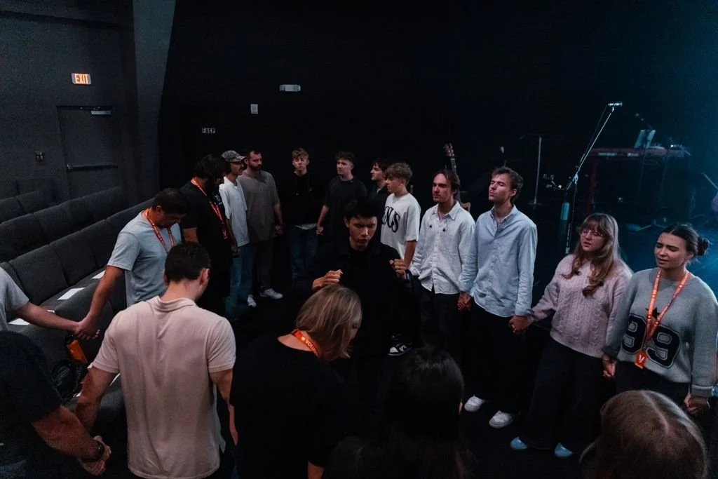 Group of people standing in a circle holding hands in a dark room, likely during a prayer or meditation session.