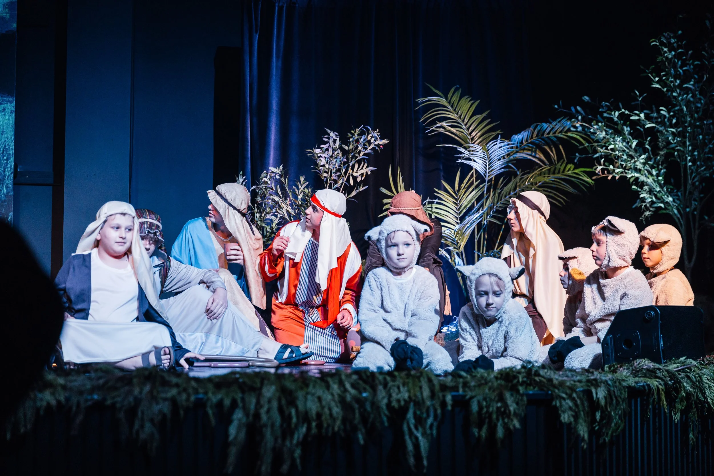 Children performing a nativity scene on stage, dressed as shepherds and animals, with trees in the background.