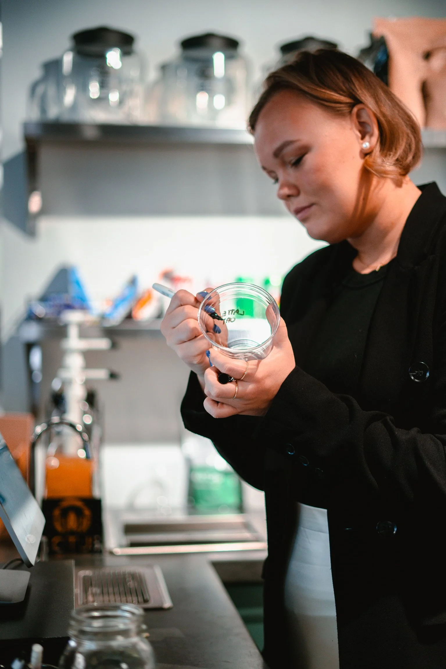 A woman in black jacket writing on a clear glass jar with a black marker in a kitchen or laboratory setting.