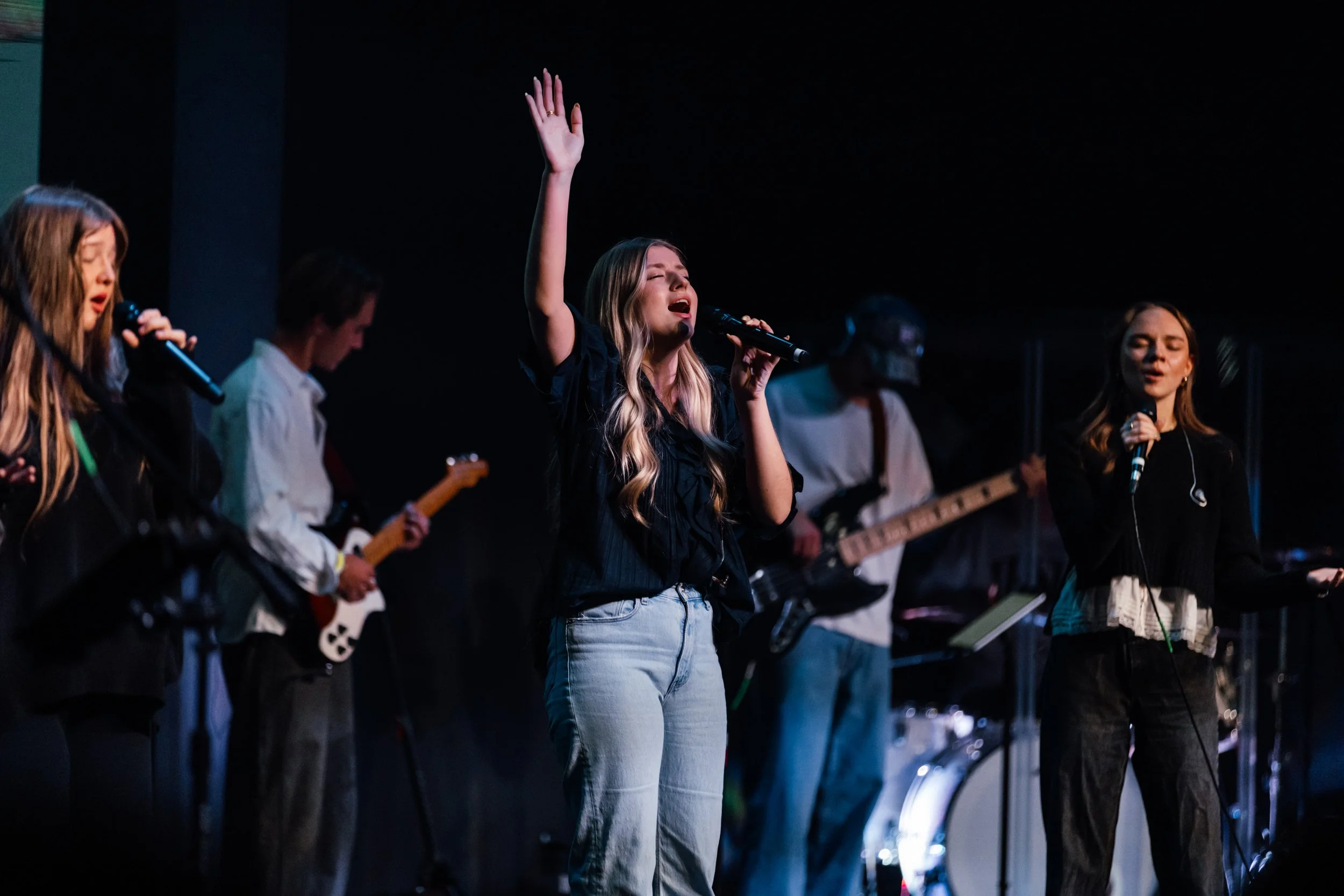 Group of young people singing and playing musical instruments on stage, with a woman in the center raising her hand, holding a microphone.