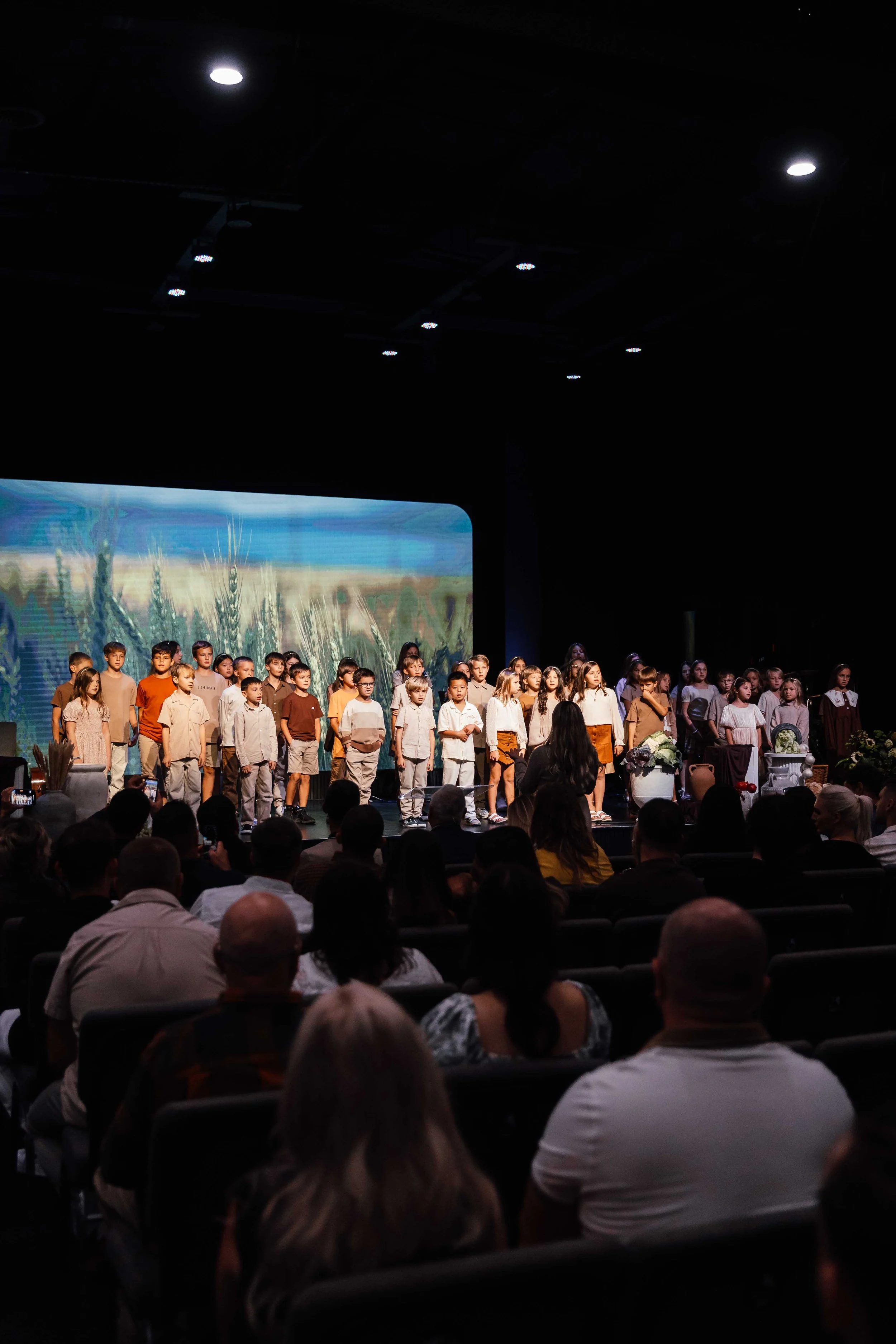 Children performing on stage in front of audience, with a nature-themed background and stage props.