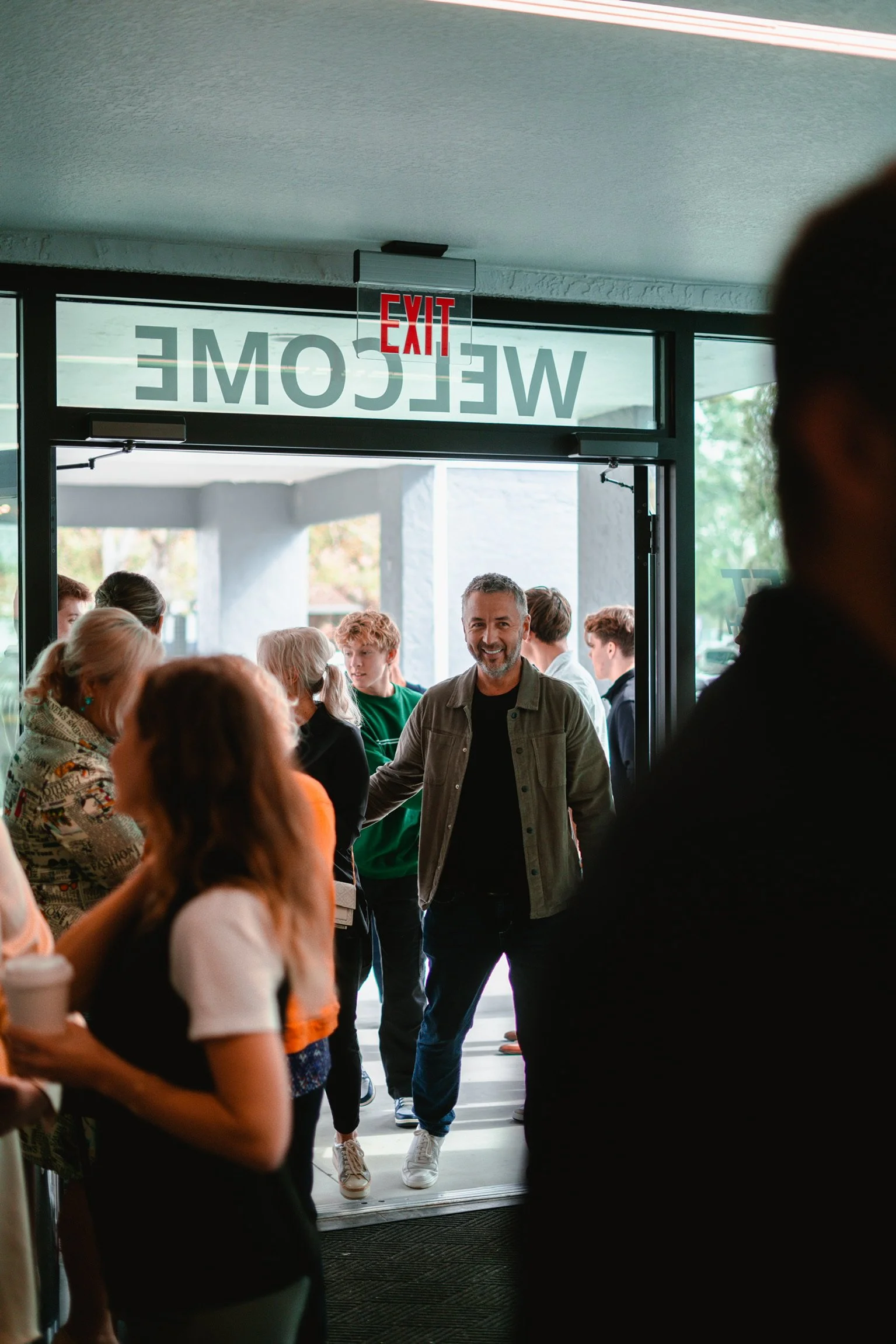 People entering a building through a glass door with an 'EXIT' sign above and a 'WELCOME' sign reflected in the glass.
