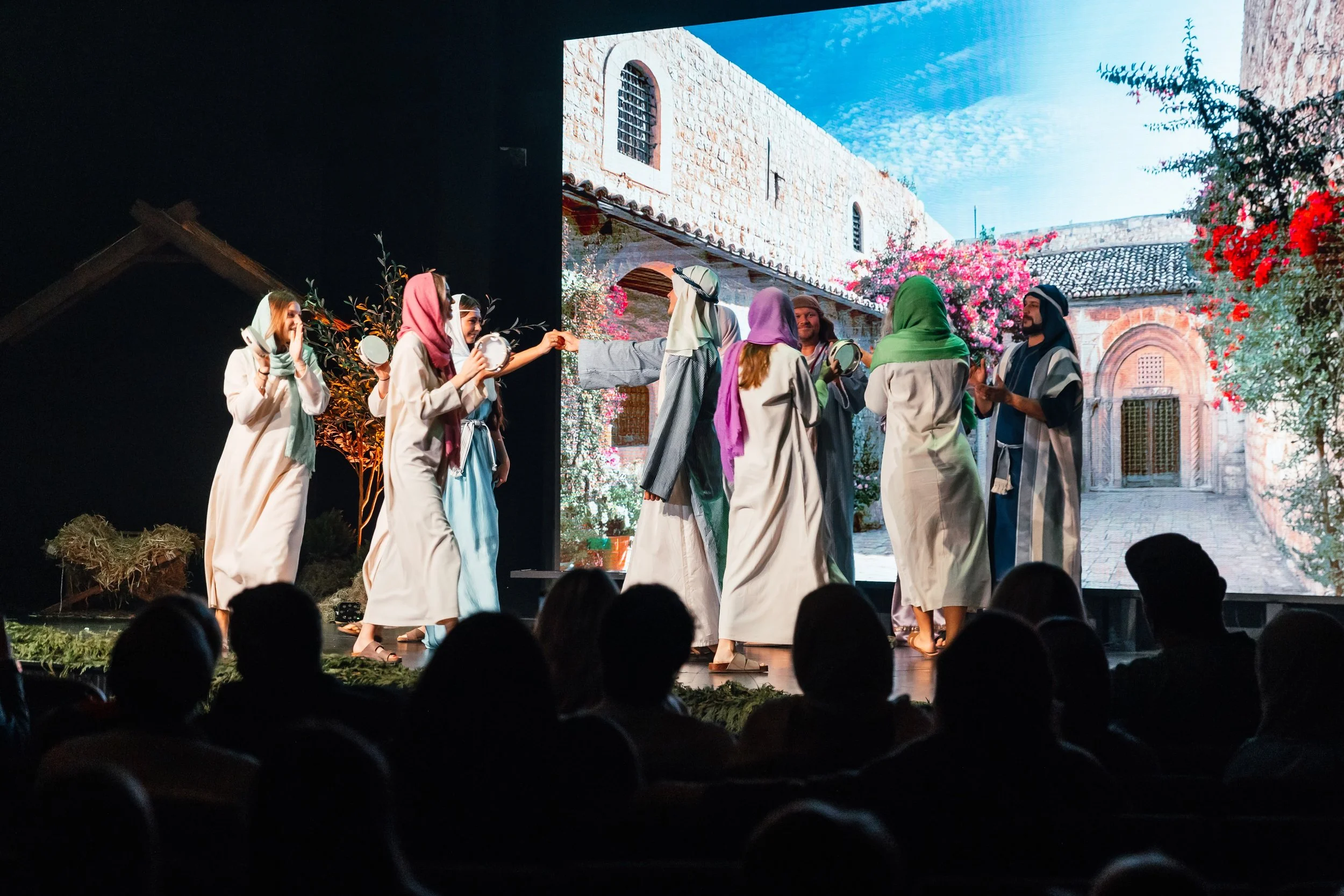 Group of performers dressed in historical clothing on stage with a scenic backdrop of a courtyard with stone buildings and flowering trees, audience watching.