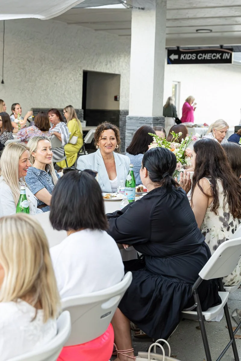 Group of women sitting at a table with flowers and drinks, engaged in conversation at a gathering or event, with a sign for kids check-in in the background.