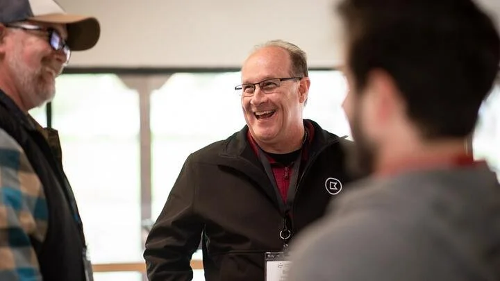 Three men talking and laughing indoors, with a large window in the background.