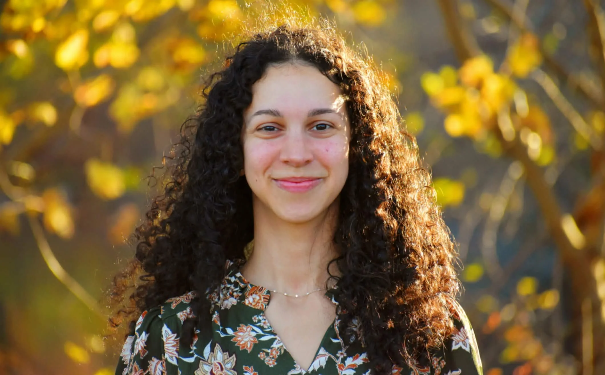 Young woman with long curly hair smiling outdoors in fall, wearing a patterned top with floral design.