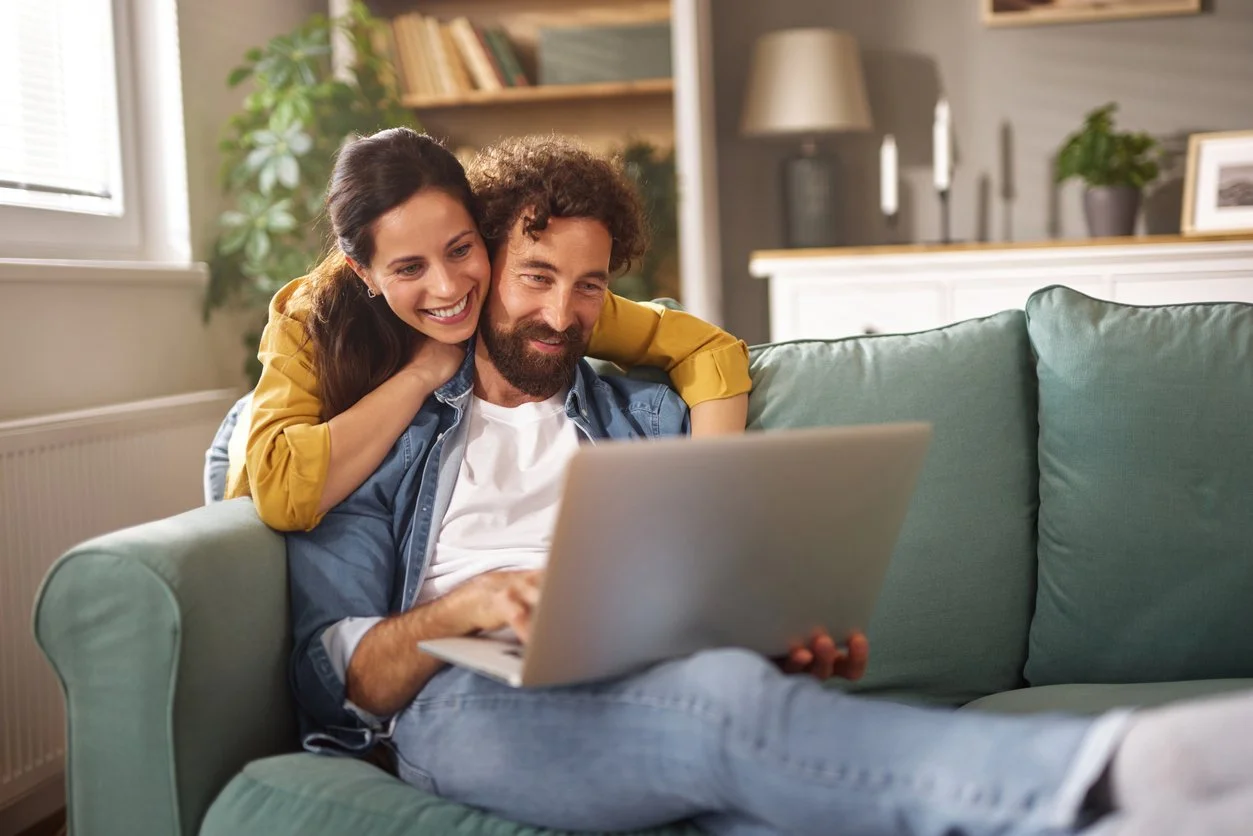 Man sitting on sofa with woman resting her arms on his shoulders, both looking at computer with soft smiles
