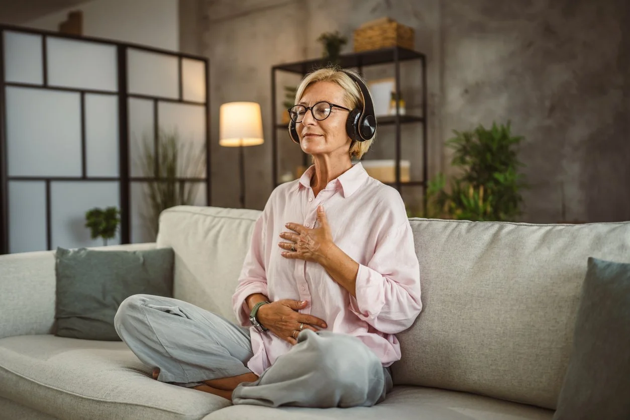 Woman peacefully sitting on sofa wearing headphones with eyes softly closed and hands on chest