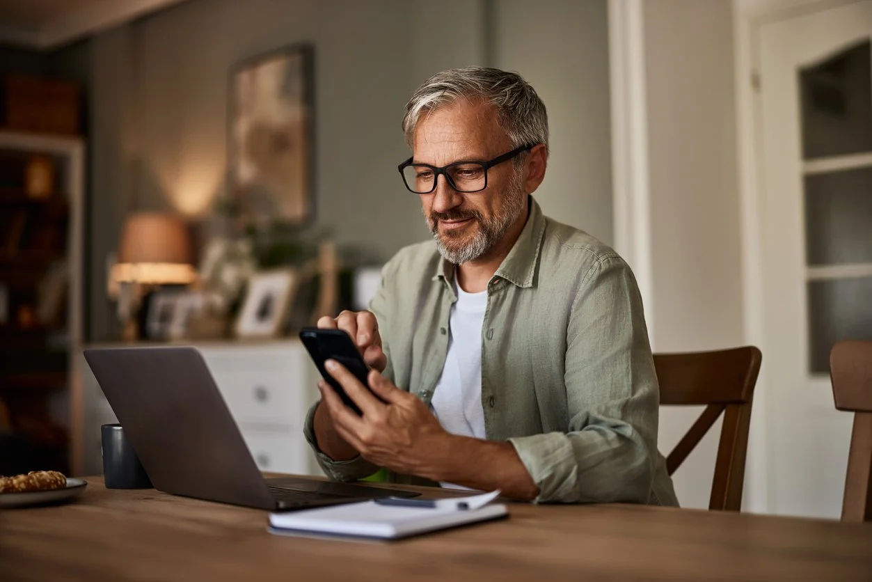Man wearing glasses typing on a smart phone with laptop on desk in front of him