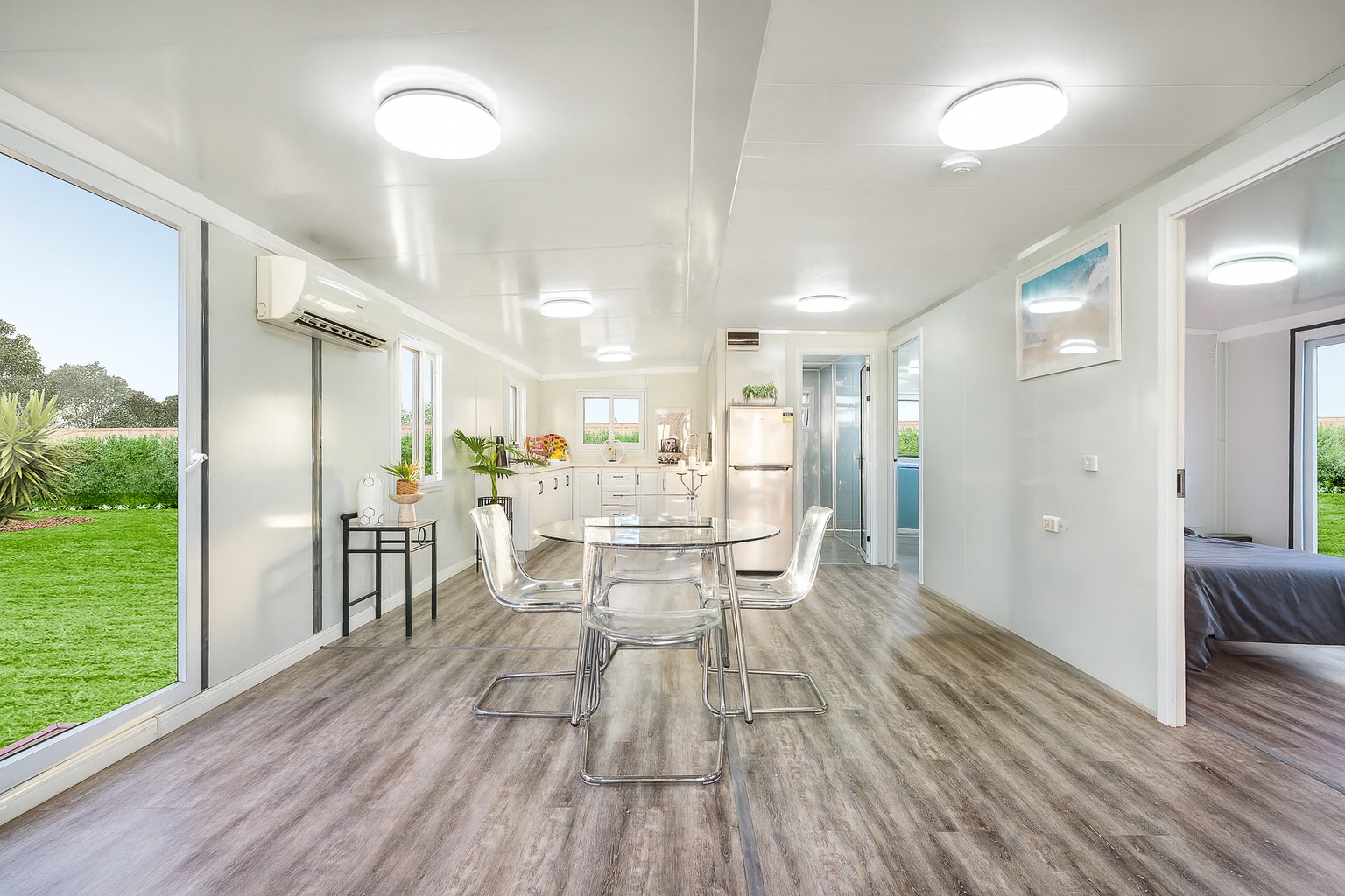 Open-plan dining area with a round glass table and four clear acrylic chairs, a kitchen with white cabinets and a refrigerator, and a bedroom visible through an open doorway, with large windows and a view of a green outdoor space.