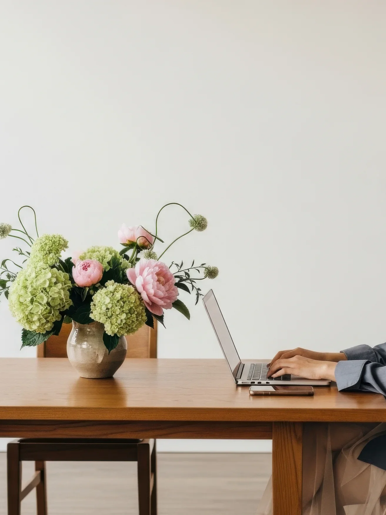 Person working on a laptop at a wooden table with a vase of pink and green flowers.