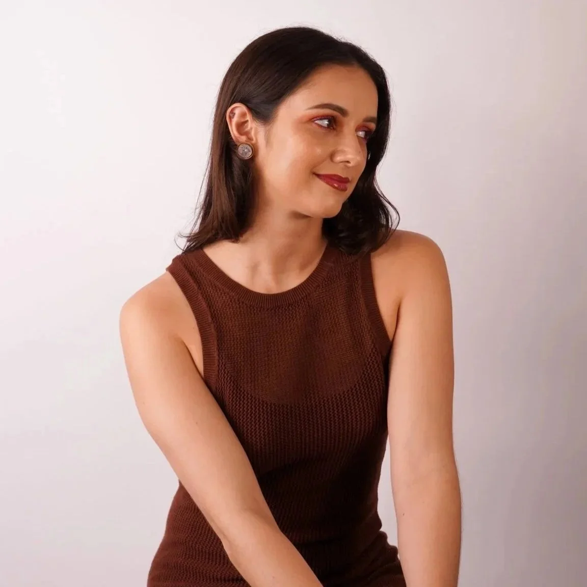 A woman with shoulder-length dark hair, wearing a sleeveless brown sweater, looking to her right with a slight smile, standing against a plain white background.
