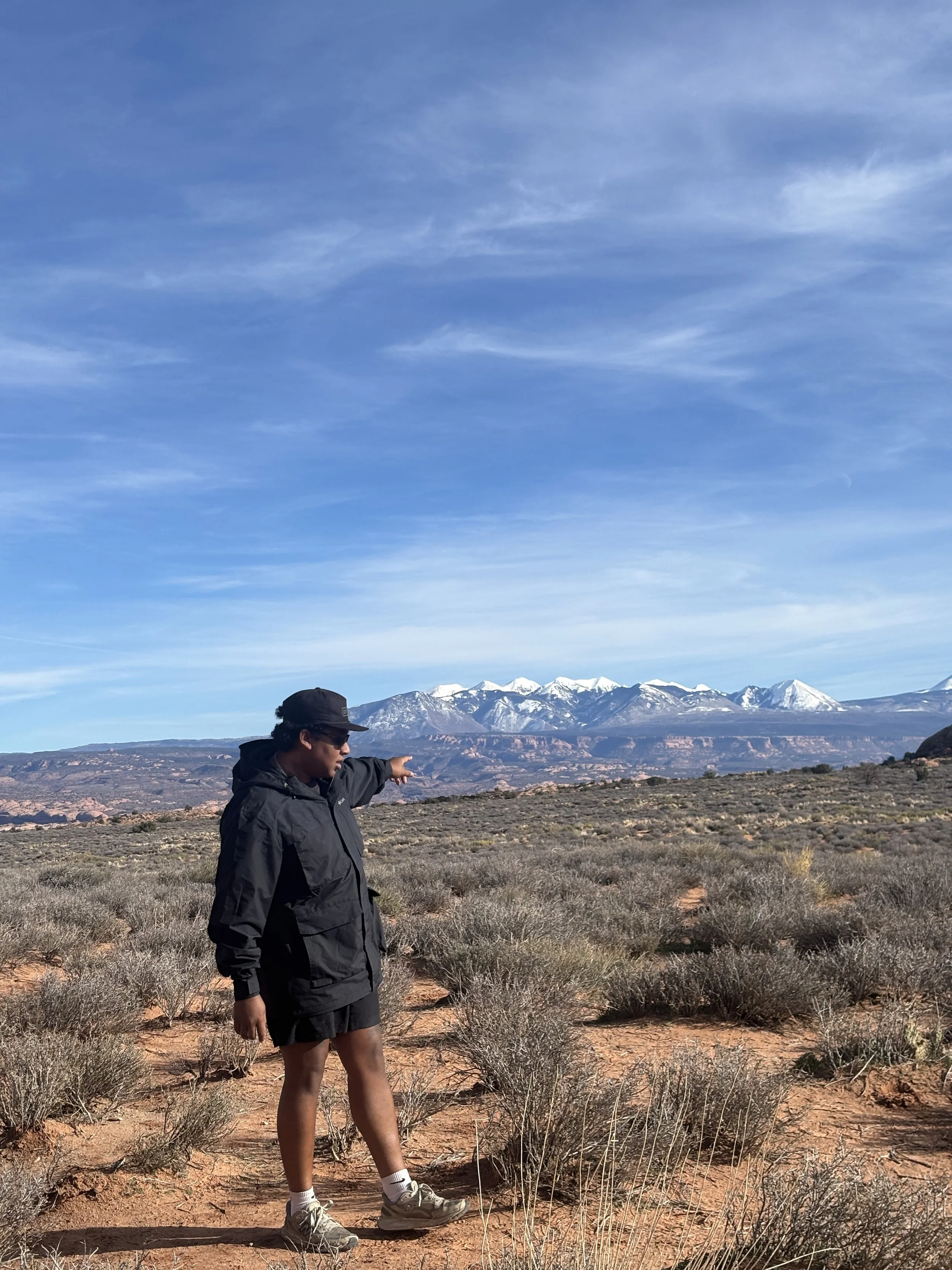 A person wearing a black jacket, shorts, and a baseball cap standing in a desert landscape pointing towards snow-capped mountains in the distance under a blue sky.