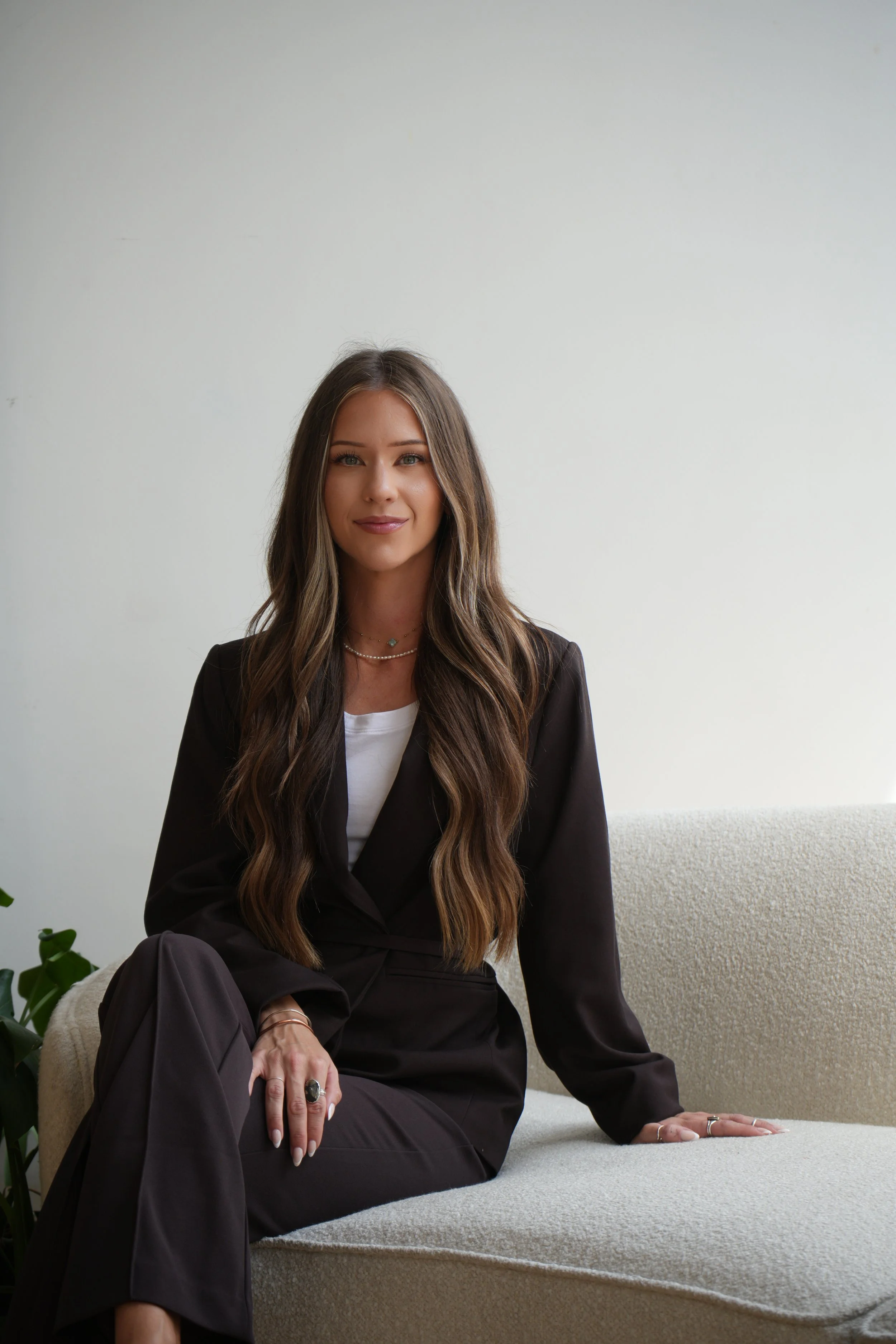 A young woman with long wavy brown hair, wearing a black blazer, white top, and dark pants, sitting on a beige sofa in a room with a plain white wall background. She has jewelry on her fingers and neck, and is looking at the camera with a slight smile.
