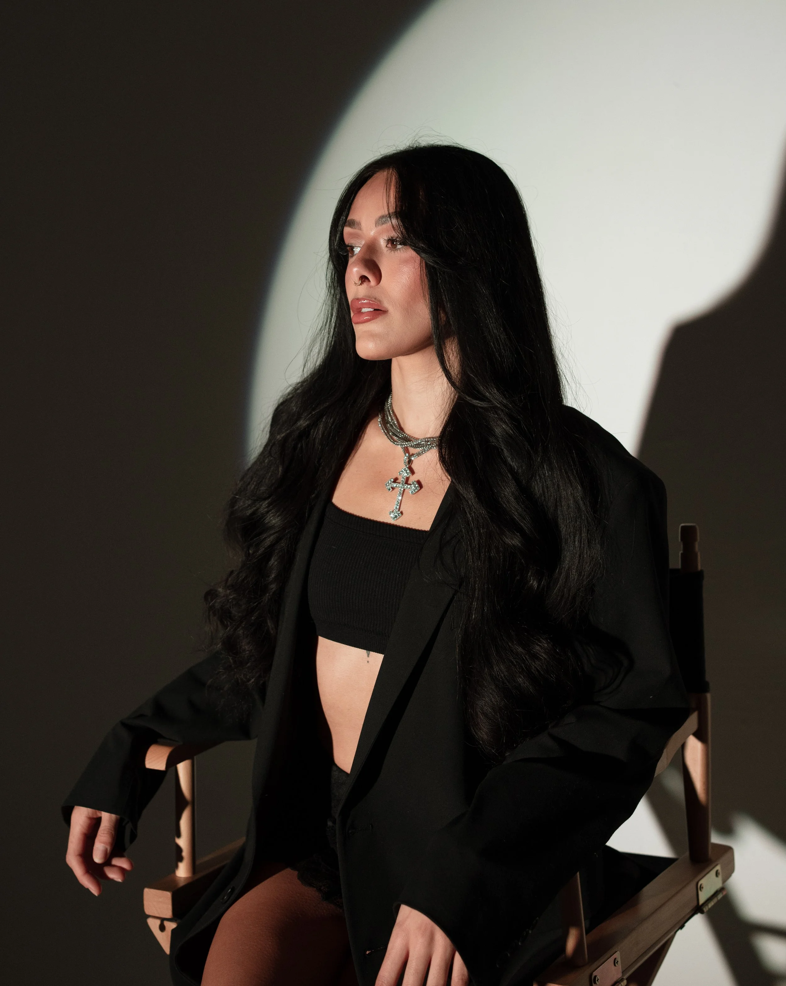 A woman with long black hair and light makeup sitting on a director's chair, wearing a black crop top, black blazer, layered necklaces with a cross pendant, and dark shorts, with a shadow cast on the wall behind her.