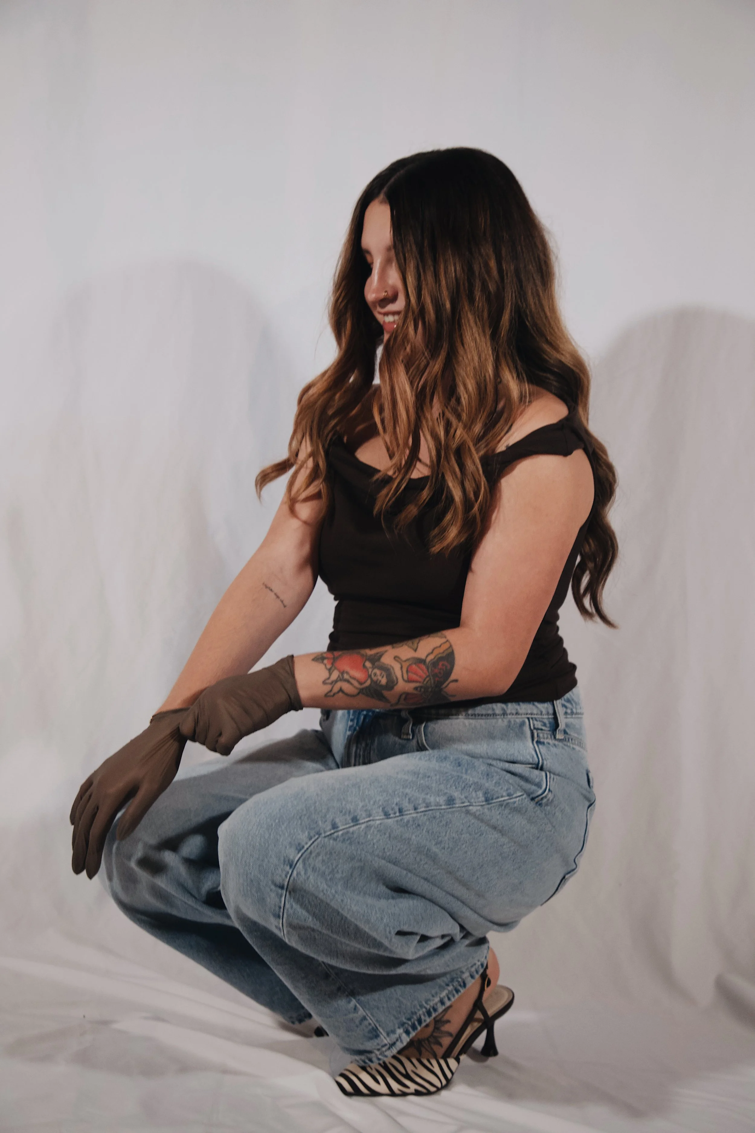 A woman with long, styled brown hair wearing a black top, light blue jeans, sunglasses, and high heels, crouching and wearing brown gloves, against a plain white background.