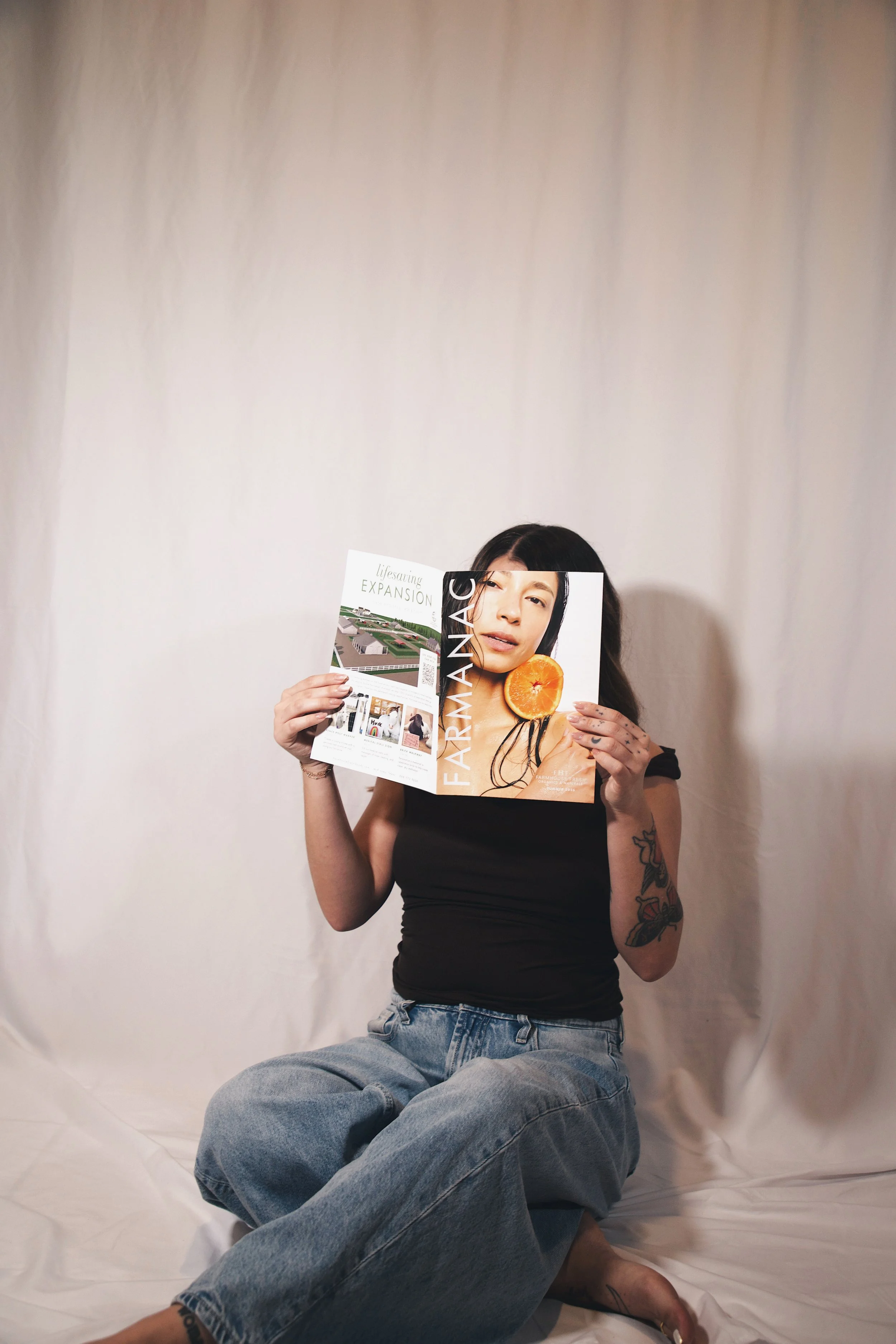 A woman with tattoos holding an open magazine, sitting on the floor with crossed legs, against a plain beige backdrop.