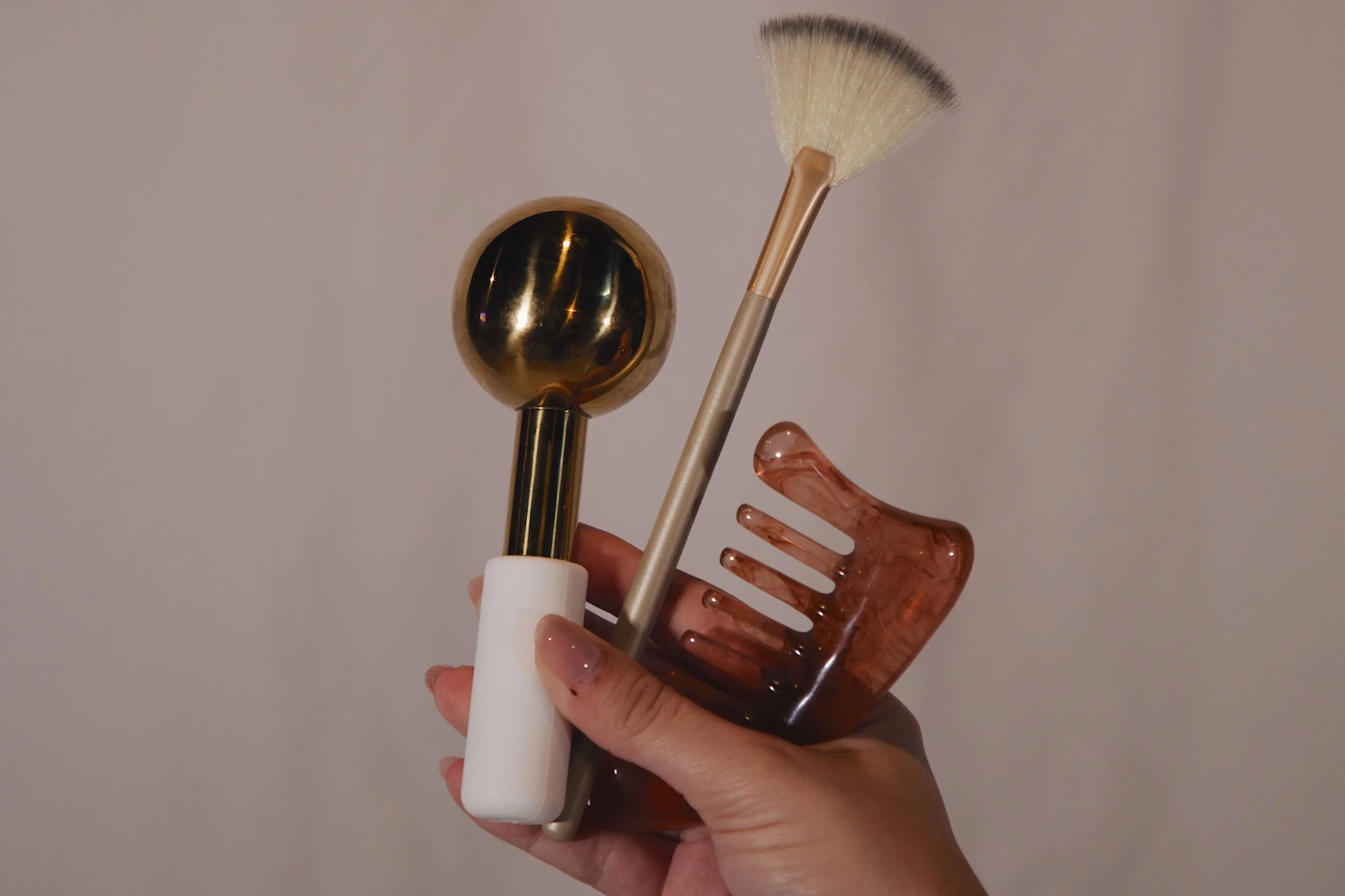 Hand holding a collection of beauty tools: a gold facial roller, a fan-shaped makeup brush, a small comb, and a pink-tinted facial massager.