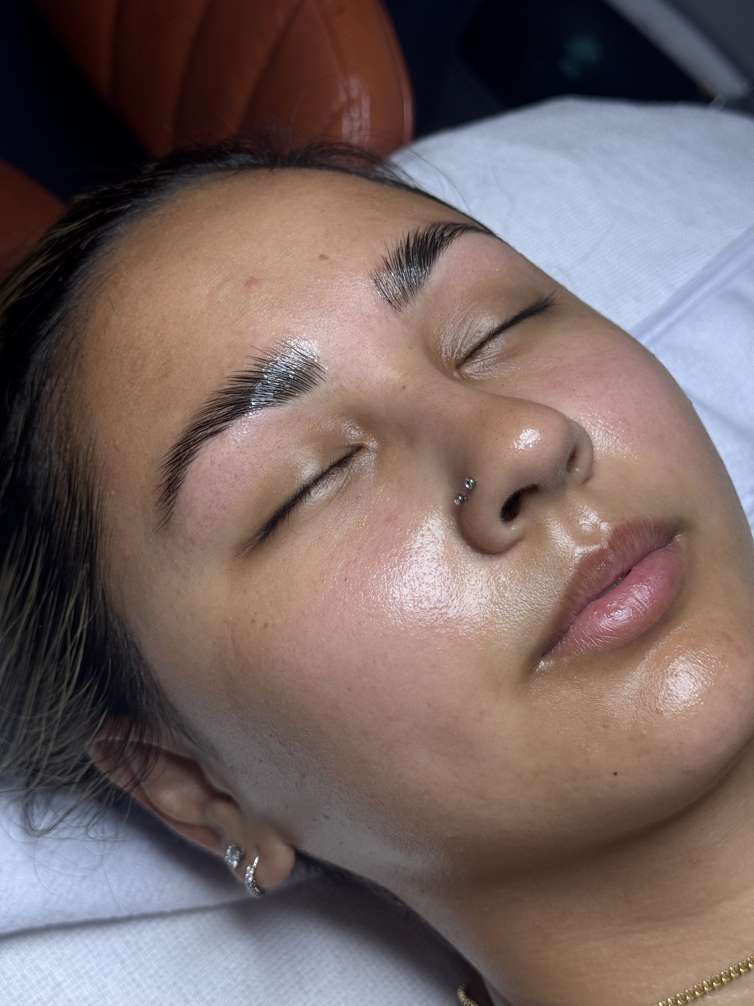 Close-up of a woman with long eyelash extensions, closed eyes, and a nose piercing, lying down with her head tilted to the side.