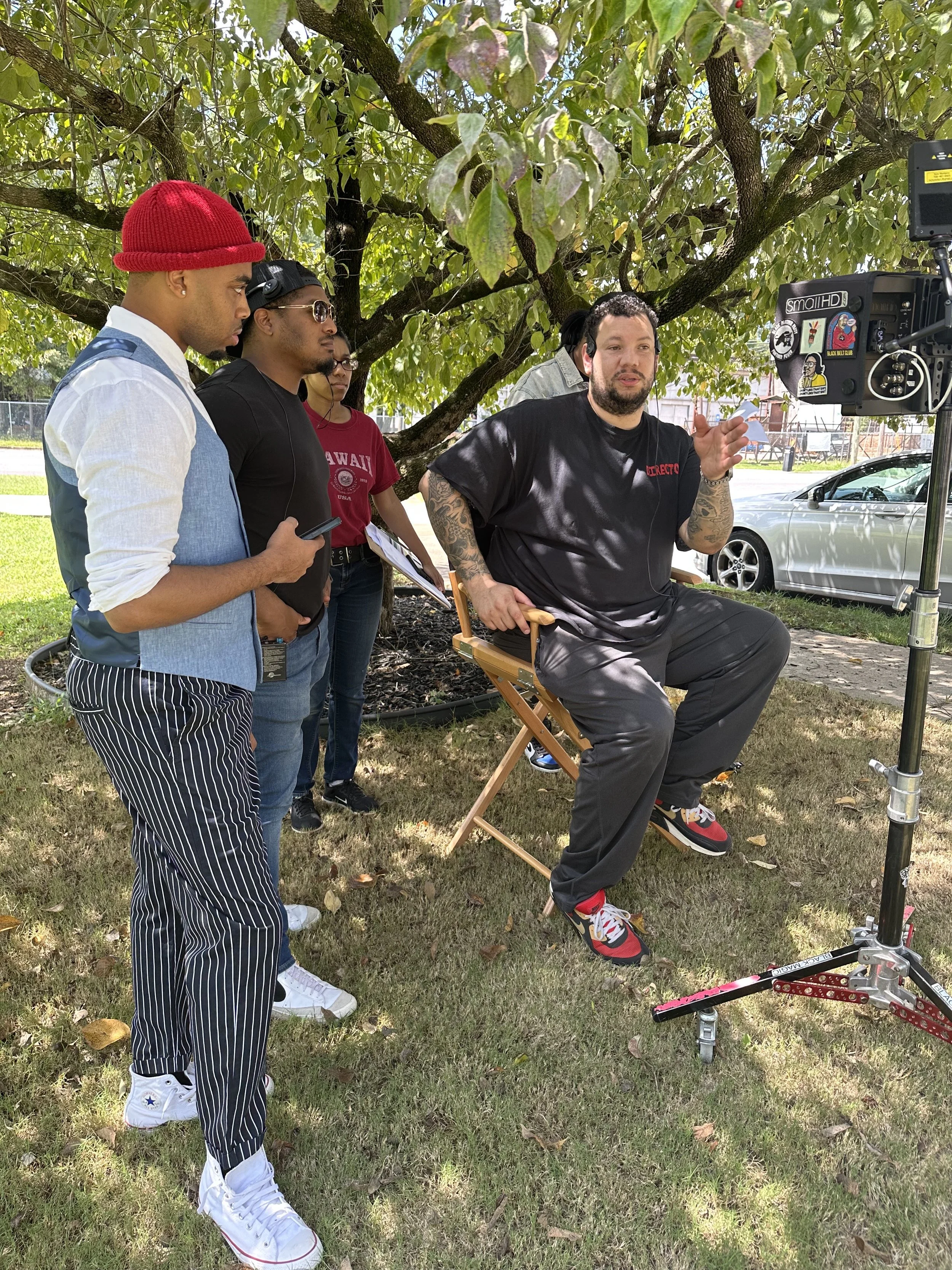 A group of four people, two men and two women, filming an interview or discussion outdoors under a large tree. One man is sitting on a director's chair, speaking into a microphone, with a professional camera on a tripod recording him. The others are standing around, listening and observing.
