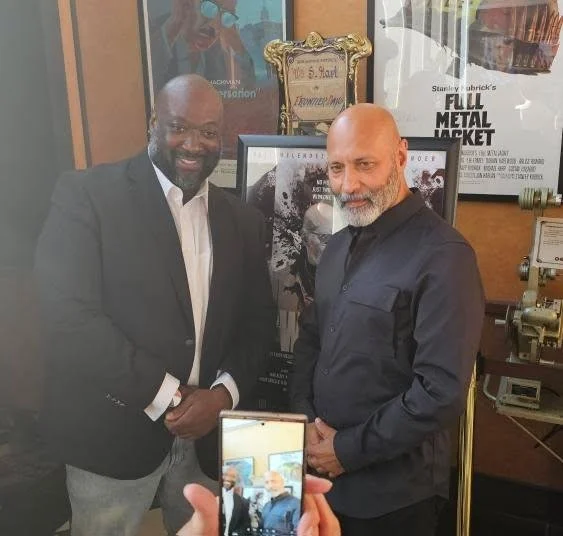 Two men in formal attire smiling and shaking hands at an indoor event, with framed pictures and posters in the background, one person taking a photo of them.