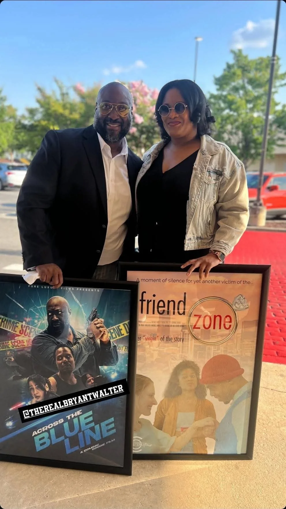Two people, a man in a black blazer and a woman in a beige jacket, standing outside next to two framed posters. The posters feature promotional images for a movie titled 'Across the Blue Line' and the story 'Friend Zone.' They are smiling and posing 