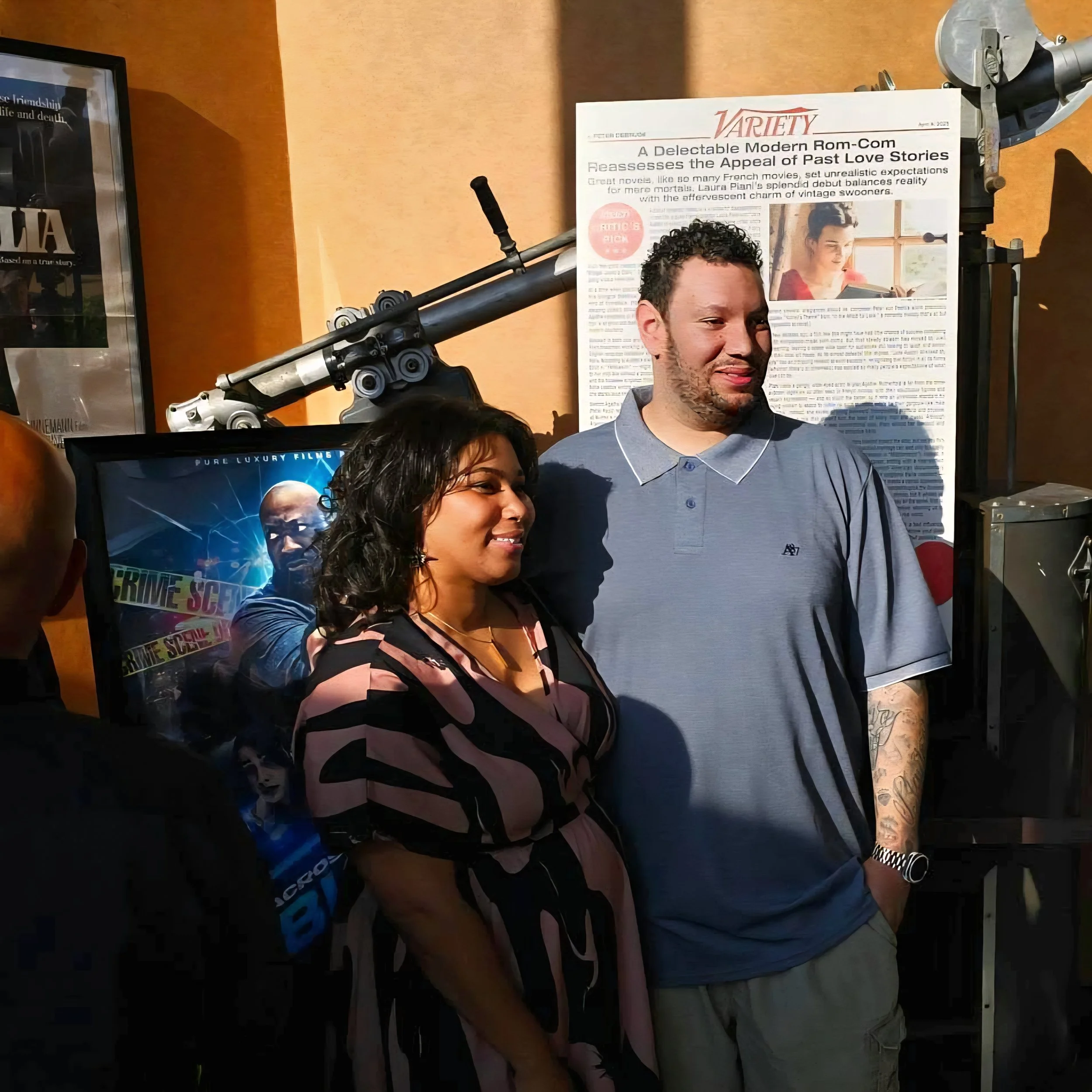 A man and woman standing together, smiling for a photo at an event. The woman is wearing a colorful, patterned dress and the man is in a gray polo shirt. Behind them are posters and a robotic arm on display.