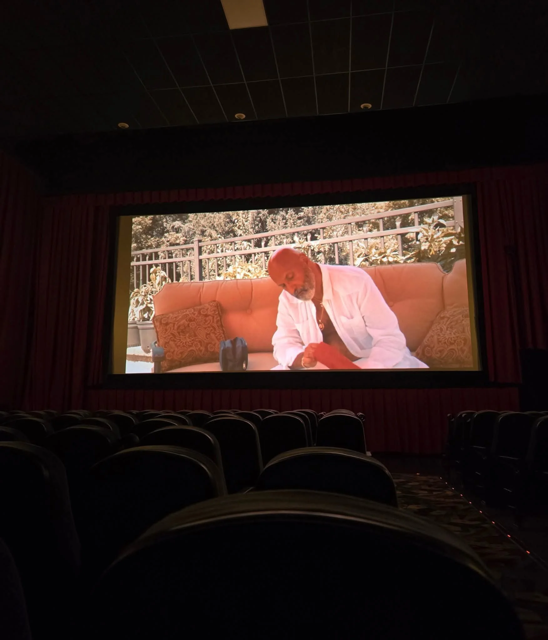 The image shows a movie theater screen displaying a scene with an older man with a bald head and gray beard sitting outside on a cushioned lounge chair, wearing a white shirt and reading a book. The theater seats are visible in the darkened foregroun