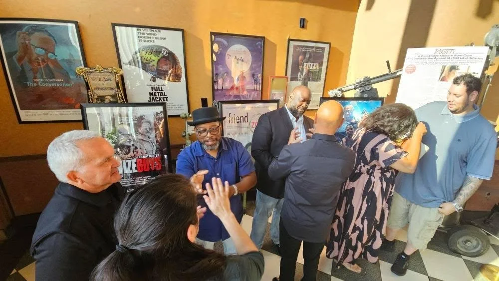Group of people engaged in conversation at an indoor event, with framed movie posters on the wall behind them.