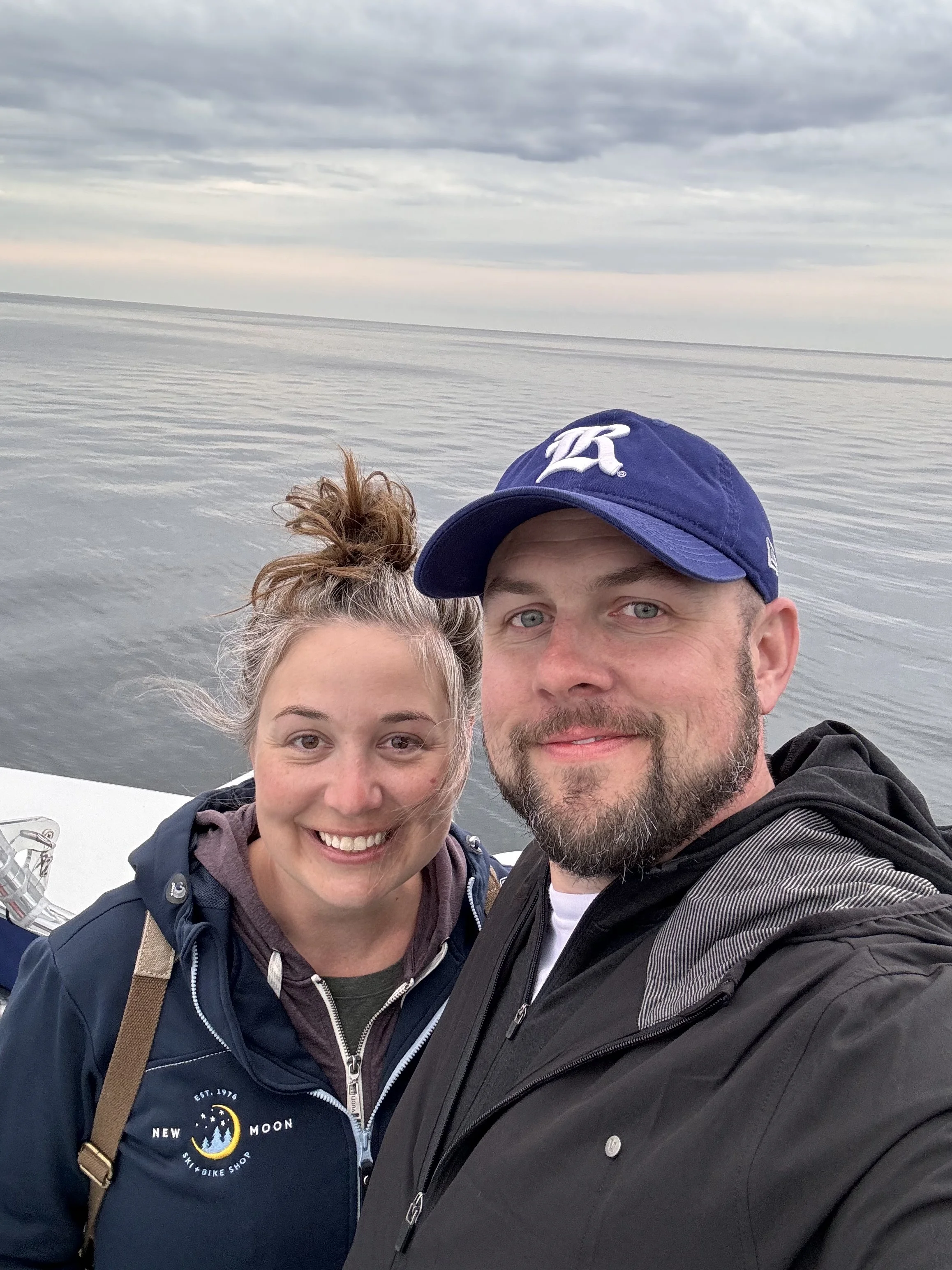A smiling couple taking a selfie on a boat with a calm body of water and cloudy sky in the background.