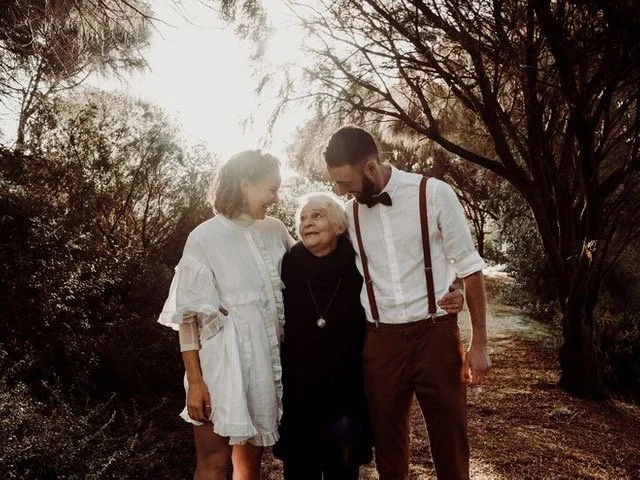 Three people, a young woman, an elderly woman, and a young man, standing outdoors under a tree, smiling and interacting close together.