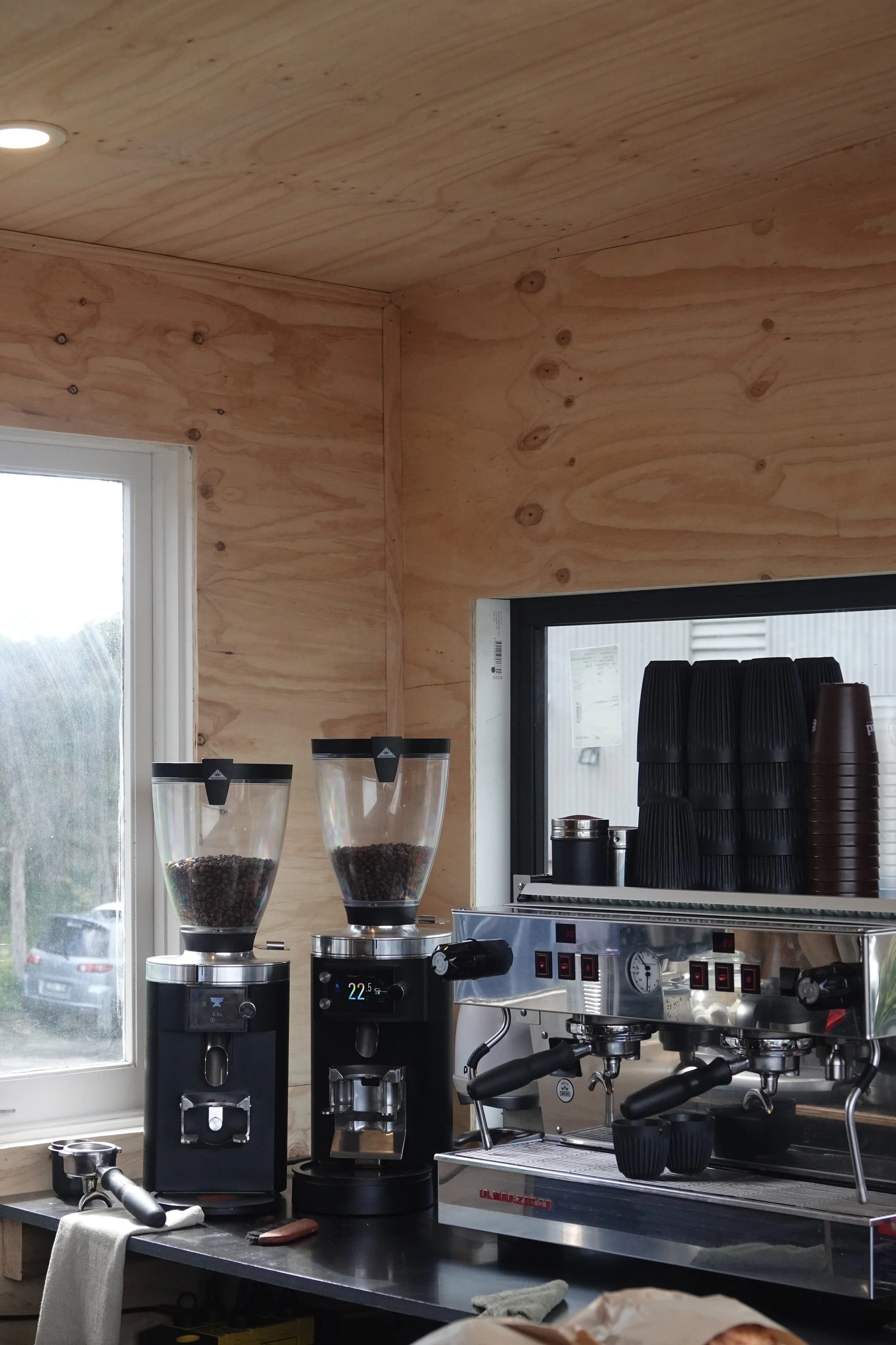 Coffee station with grinders and espresso machine, black cups stacked on top, in a room with wooden walls and a window