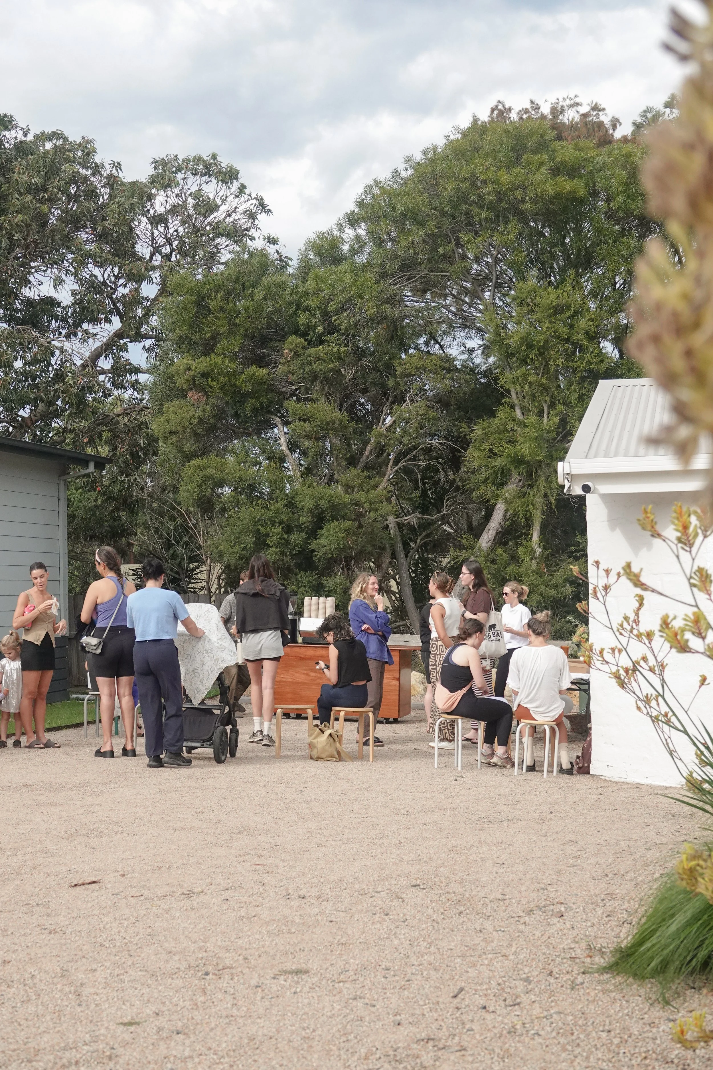 People gathered outdoors around tables with a backdrop of trees and a building, under a cloudy sky.