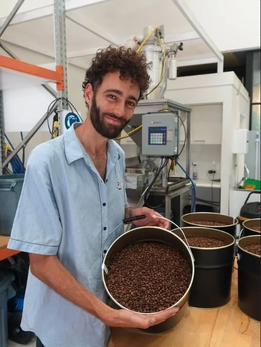 A man with curly hair and a beard holding a bucket of roasted coffee beans in a lab or industrial setting with electronic equipment and containers of coffee beans on the table.