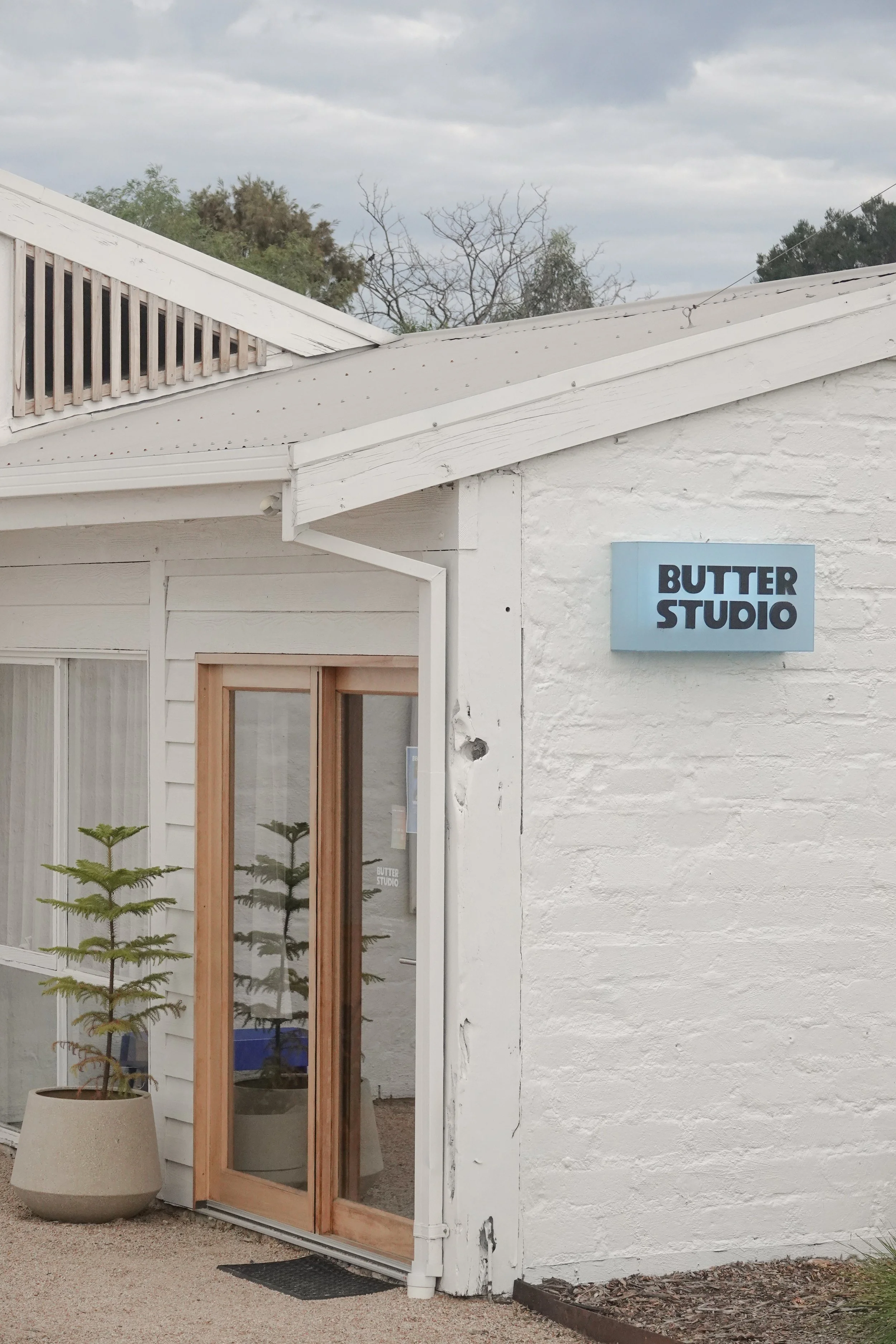The exterior of a building with a sign that reads 'Butter Studio.' The building has white walls, a wooden-framed glass door, a potted plant outside, and trees in the background under a cloudy sky.
