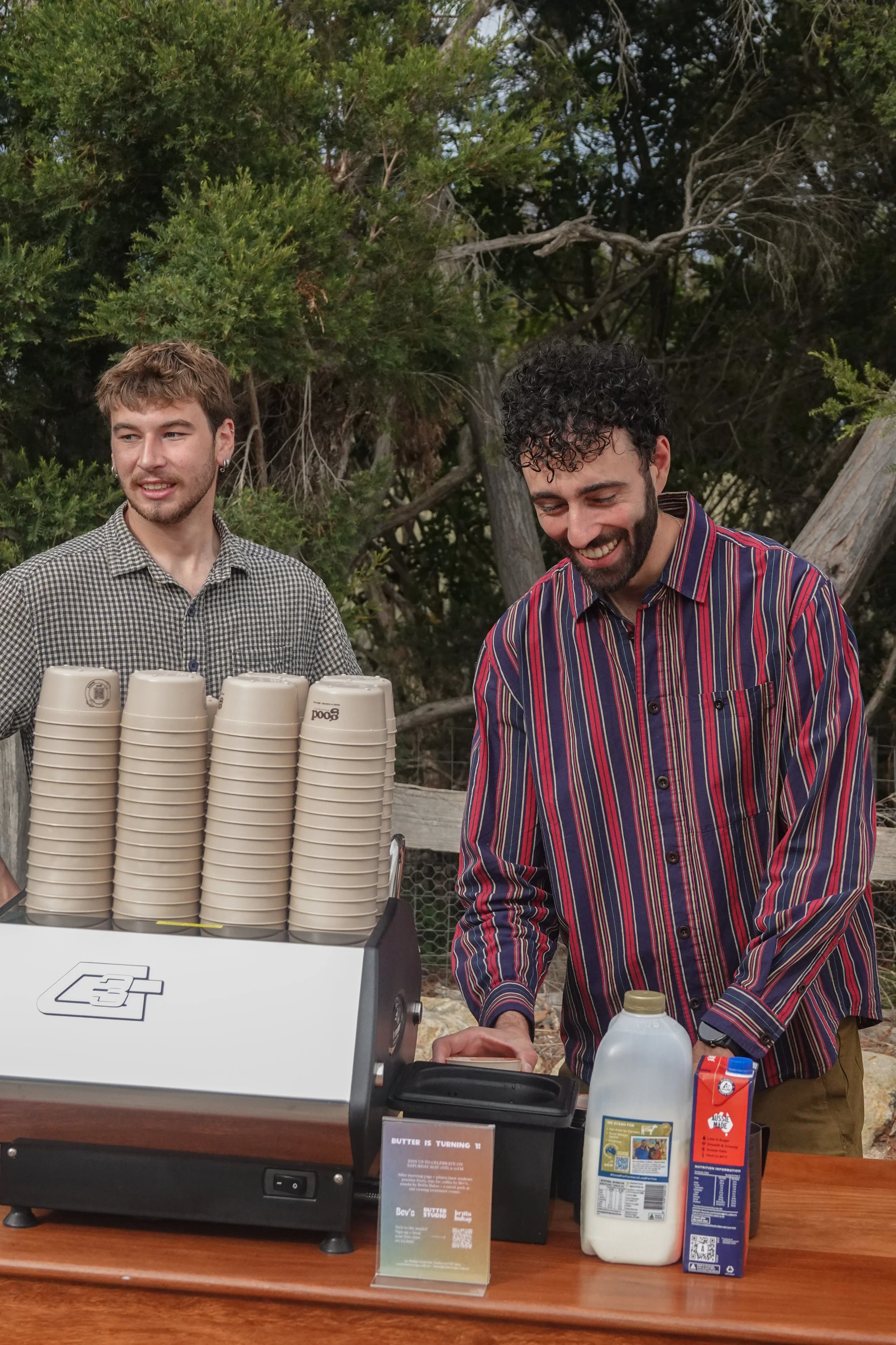 Two young men are standing behind a counter with a stack of disposable cups and various condiments. One man, wearing a checkered shirt, is looking to the side, while the other, wearing a striped shirt, is smiling and looking down. There are trees in the background.