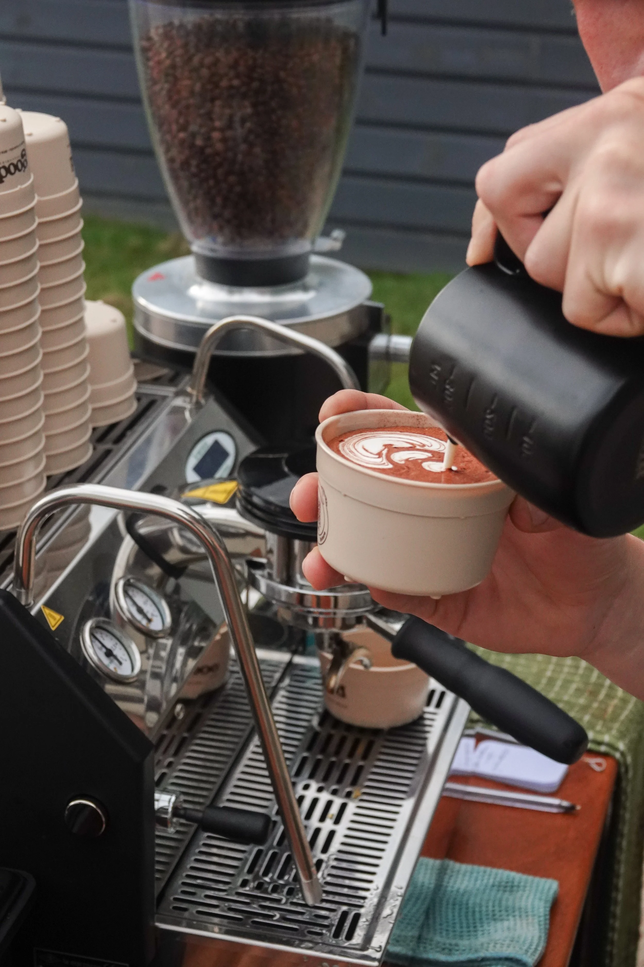 A person is pouring steamed milk into a cup of espresso to make a latte. The scene takes place on a professional coffee machine outside, with the person holding the cup with one hand and pouring with the other.