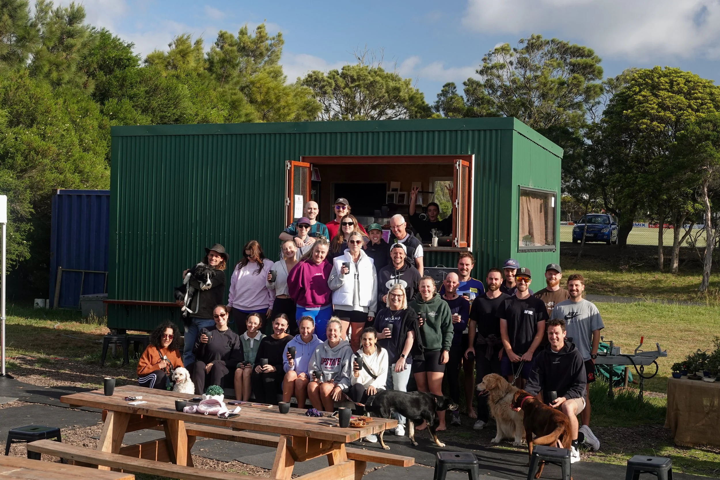 A group of people and dogs gathered outdoors in front of a green container building on a sunny day, with trees and a car in the background.