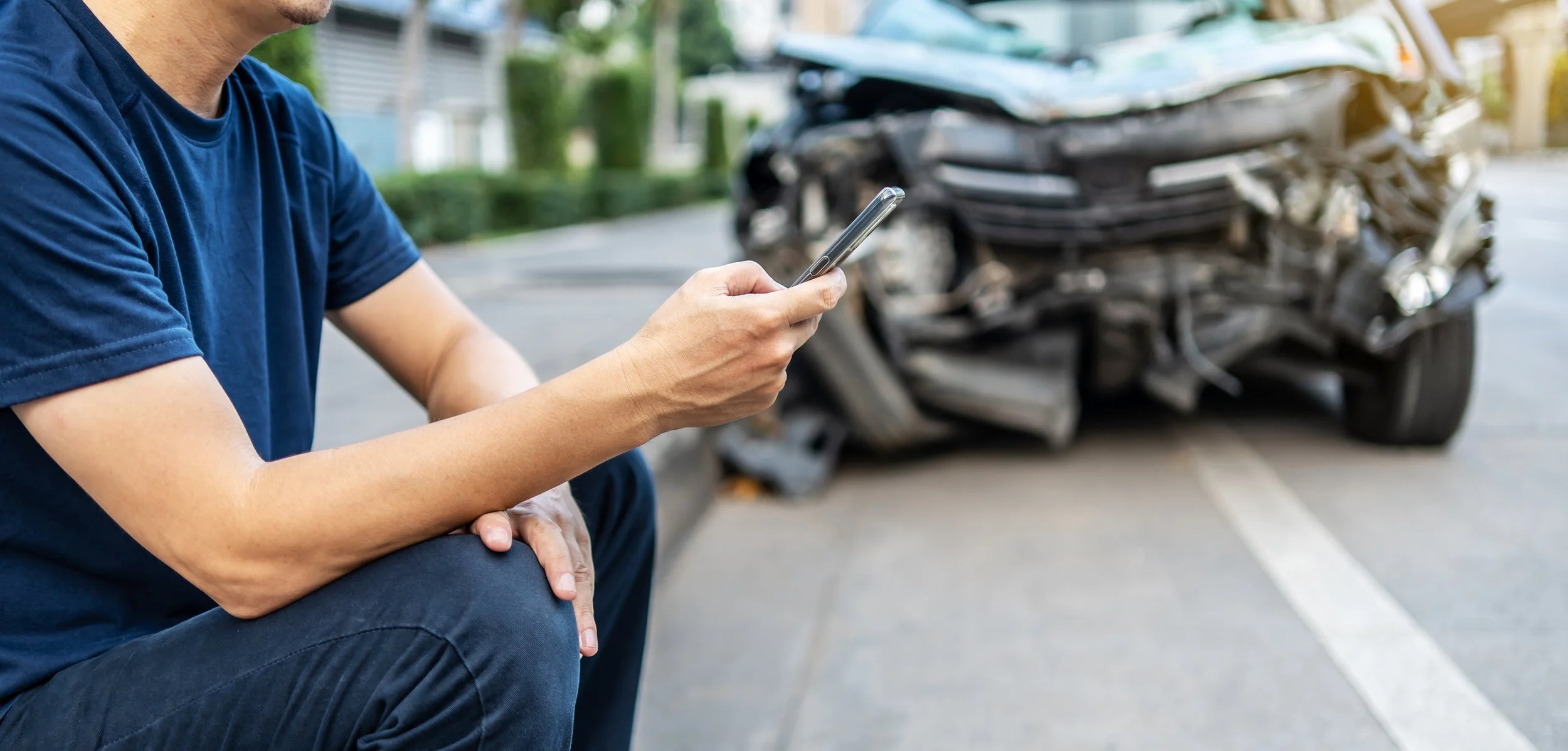 Man sitting on the curb using a phone, with a heavily damaged car in a traffic accident behind him.