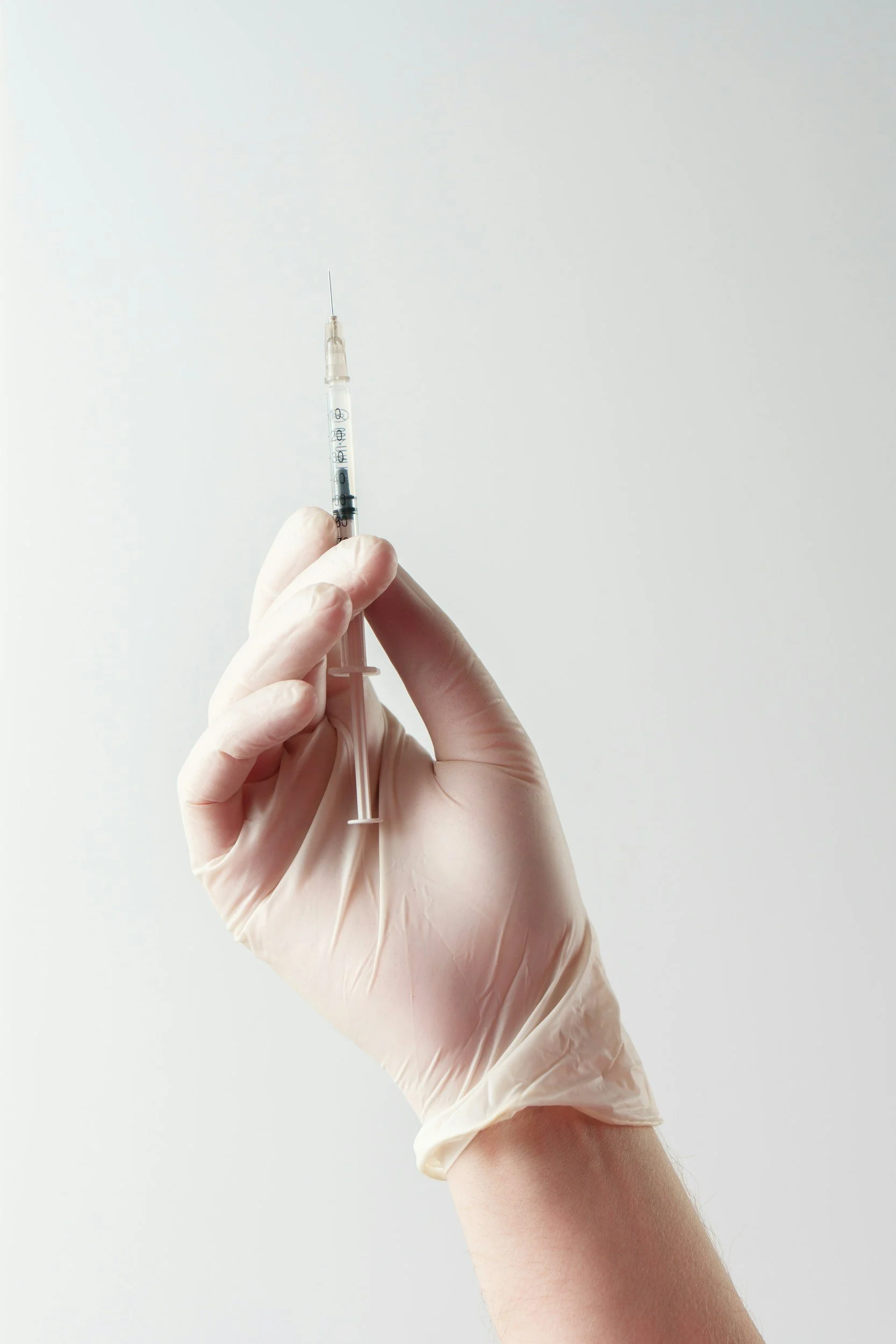 A hand wearing a latex glove holding a syringe with a needle, against a plain light background.